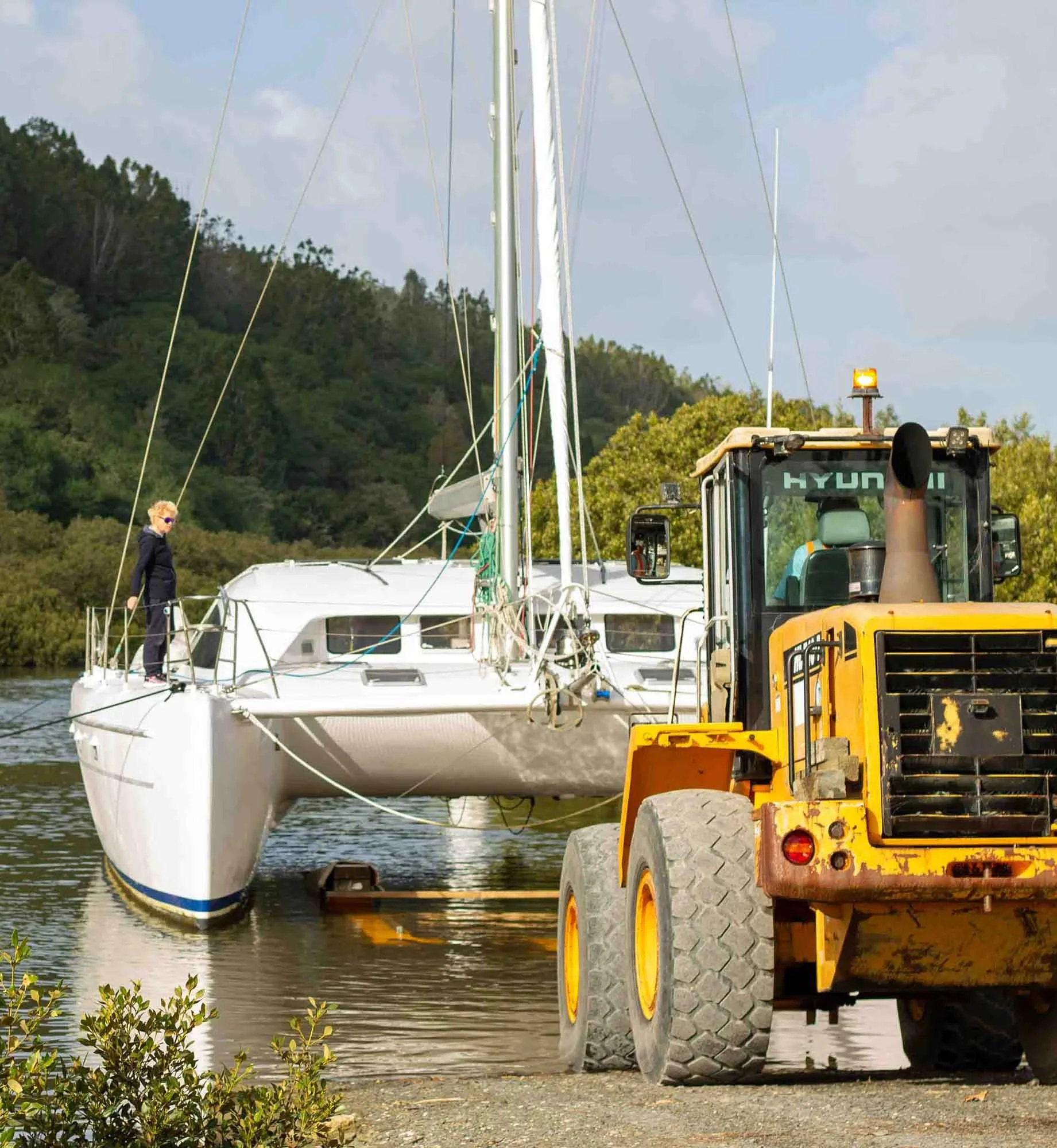 A scene of a marina with a white sailboat docked in the water and a yellow construction vehicle parked nearby on a dirt area, with green trees and hills in the background under a cloudy sky.