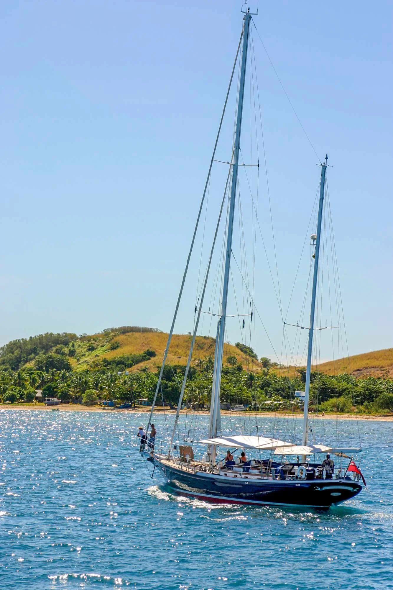 A sailboat with people on board sailing on a sunny body of water, with a green hillside and trees in the background.