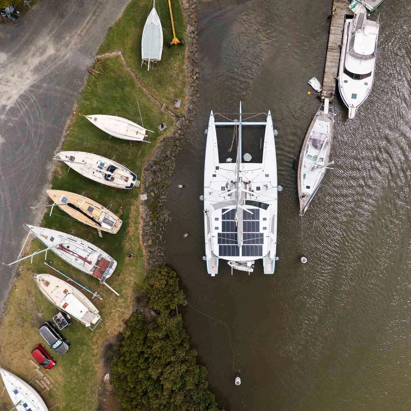 An aerial view of a marina with several boats docked and parked on land, some boats are sailboats and others motorboats. There is a grassy area with boats lying on the grass, a small parking area with cars, and a seawall with rocks. The water is calm, and the boats are secured to the docks.