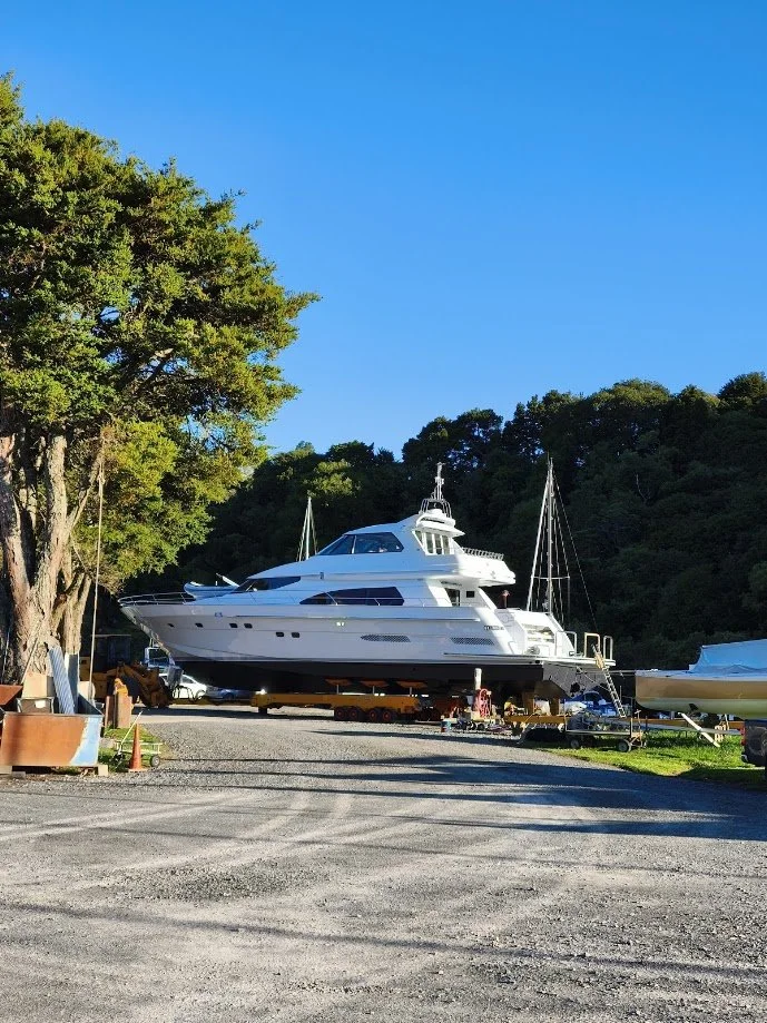A luxury yacht on land in a boatyard, surrounded by trees and equipment, with a clear blue sky.