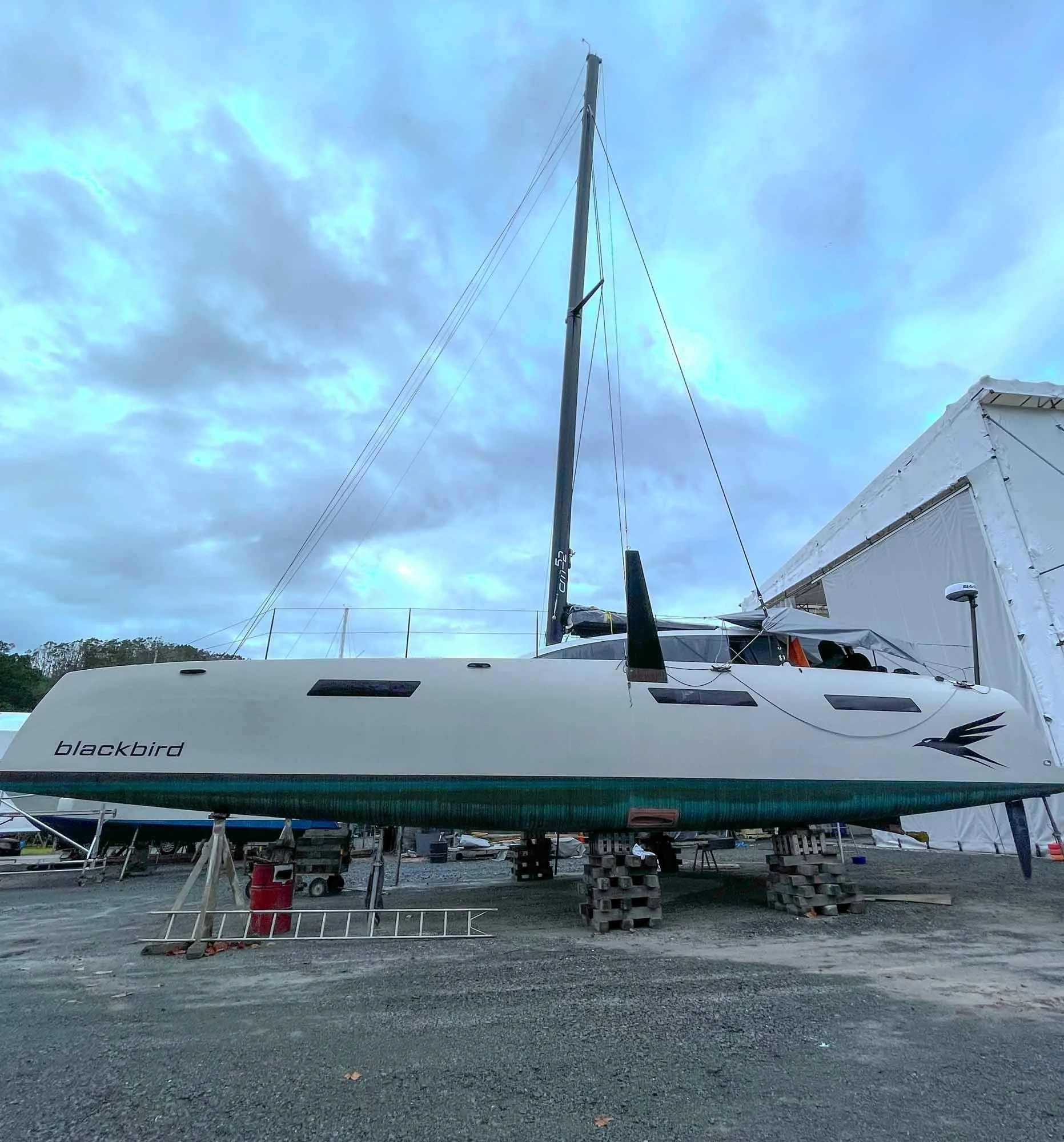 A sailboat named Blackbird on a boat trailer supported by wooden blocks at a marina, with cloudy sky overhead.