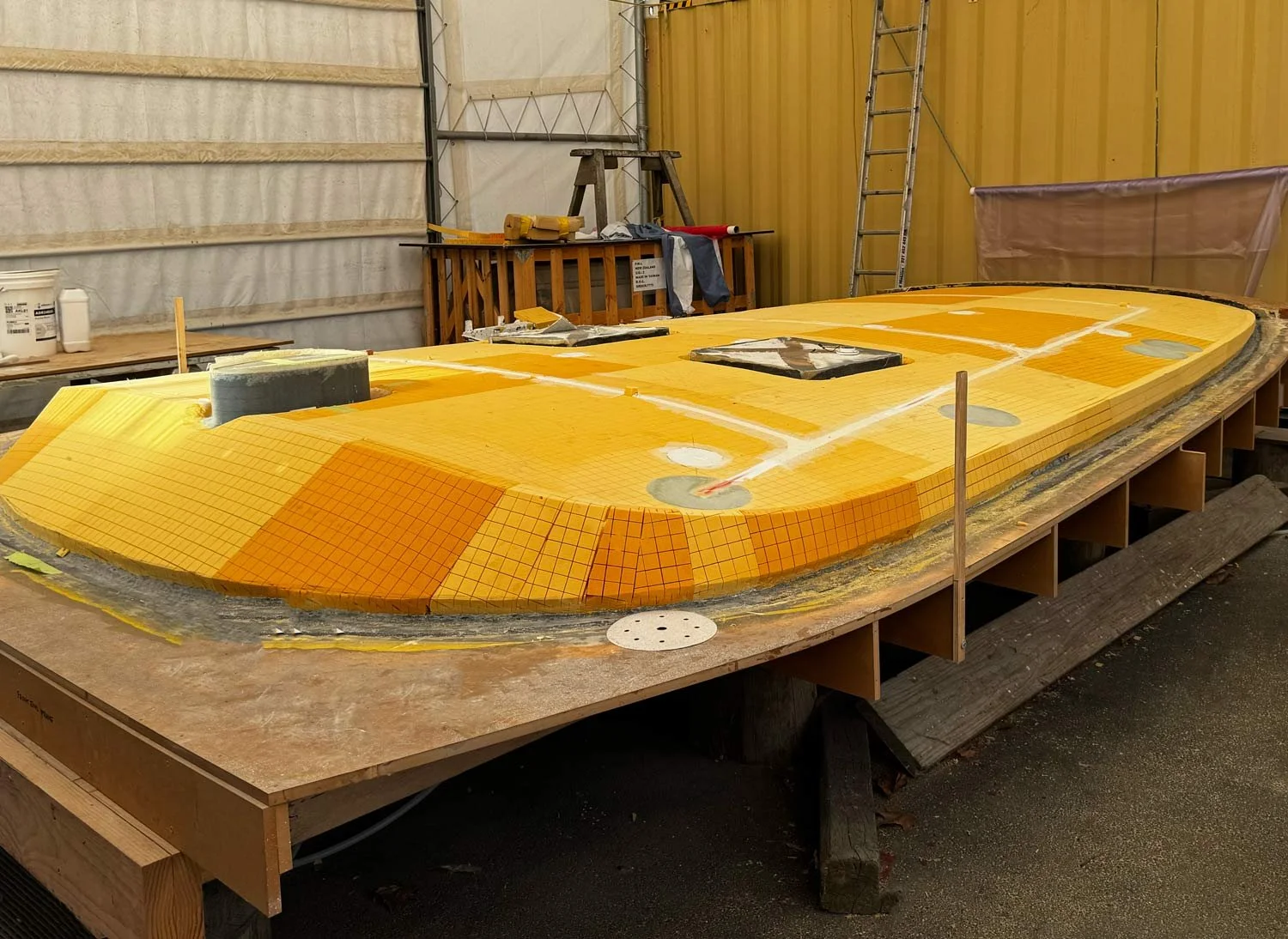 A boat hull under construction, covered in orange and yellow tiles, in a workshop setting with tools, ladder, and supplies around.