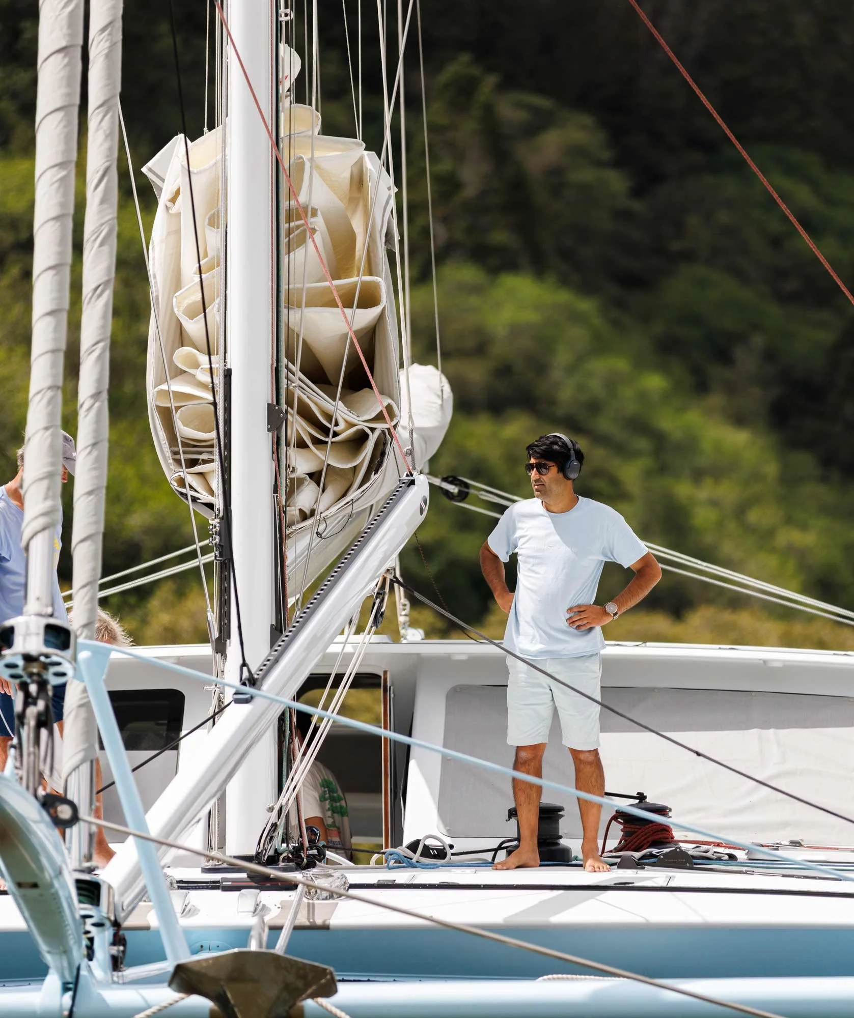 A man standing barefoot on a sailboat deck, wearing sunglasses, a white t-shirt, and white shorts, with one hand on his hip, near the sailing rig and gear, against a backdrop of green, wooded hillside.