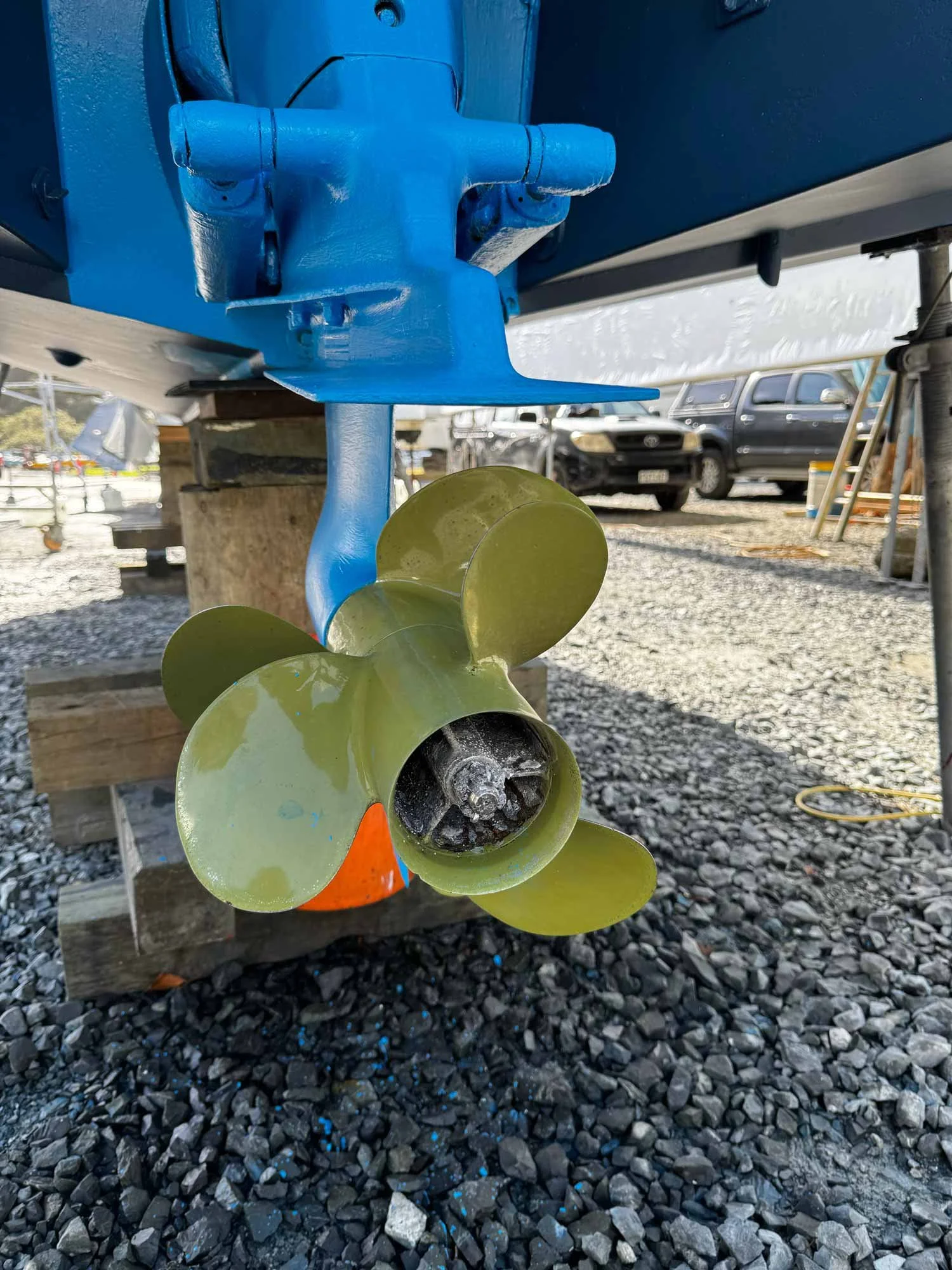 Close-up of a boat's propeller and rudder assembly mounted on a wooden support, with boats, watches, and parked cars in the background.