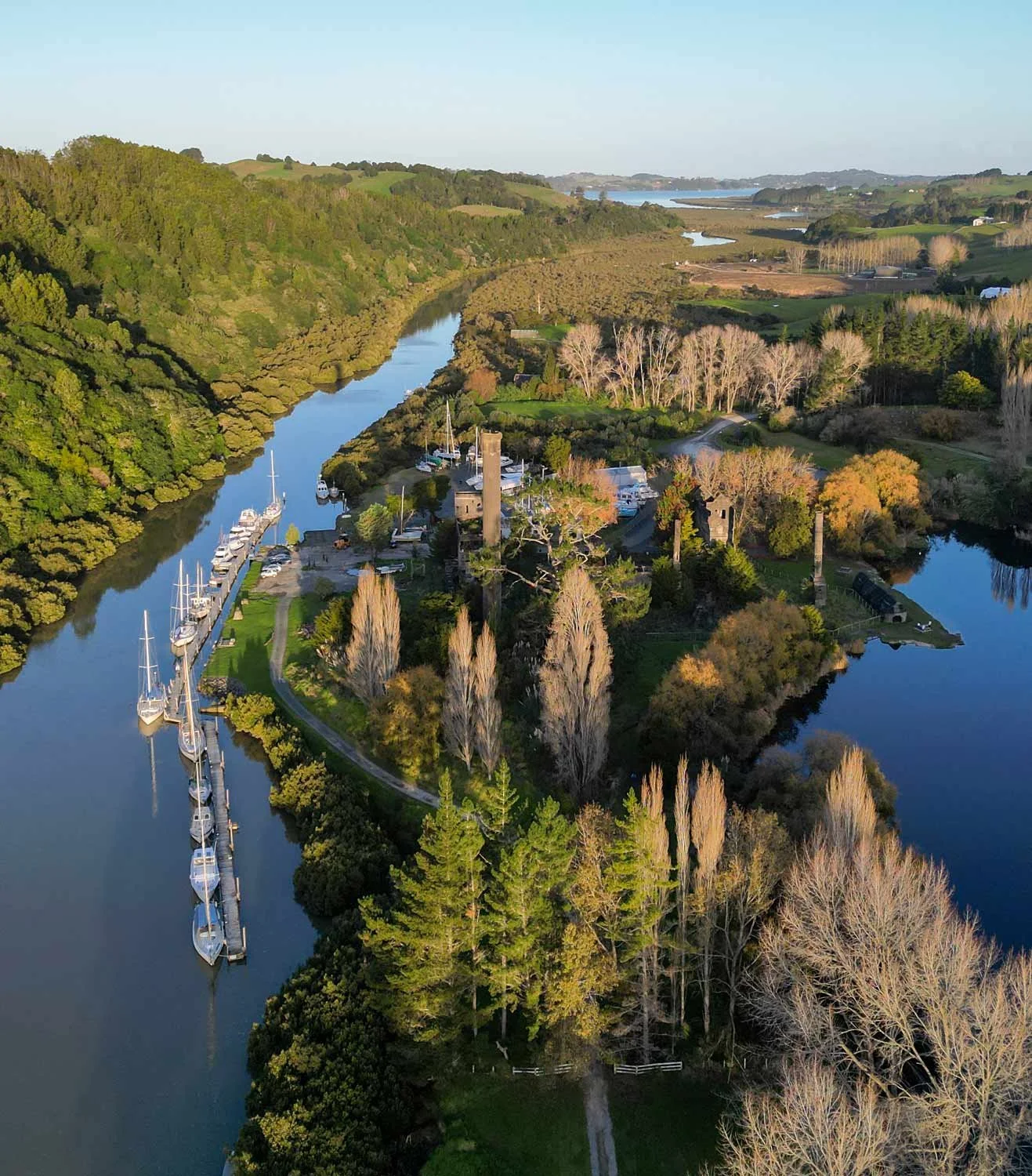 Aerial view of a marina with boats docked along a river, surrounded by lush green and autumn-colored trees, rolling hills, and distant waterways.