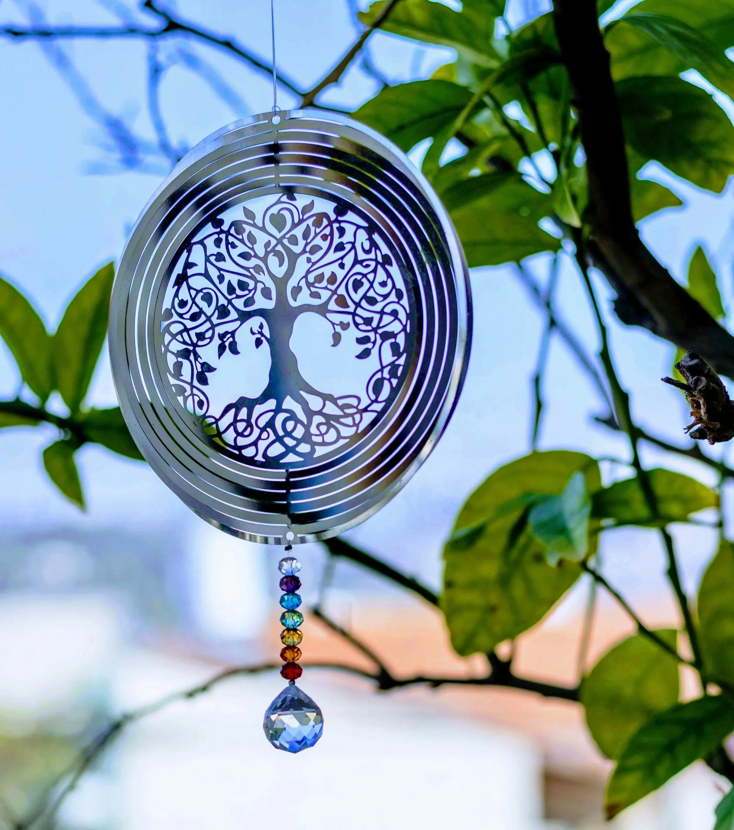 A decorative hanging ornament with a metal tree of life cutout design, beads arranged in rainbow colors according to the body's energy chakras, and a faceted crystal at the bottom, hanging among green leaves against a bright sky background.