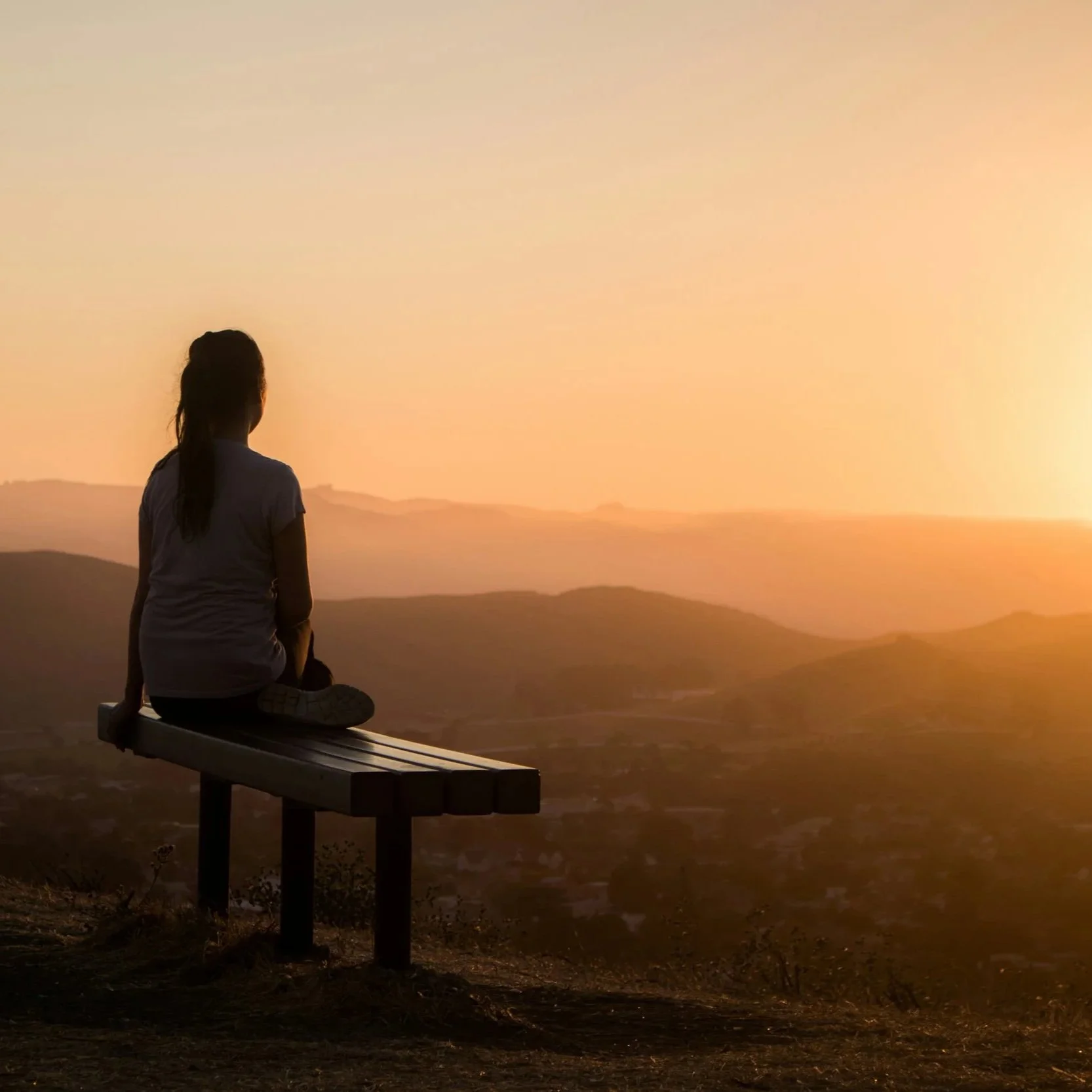 A person sitting on a park bench, watching a sunset over a landscape of hills and mountains.