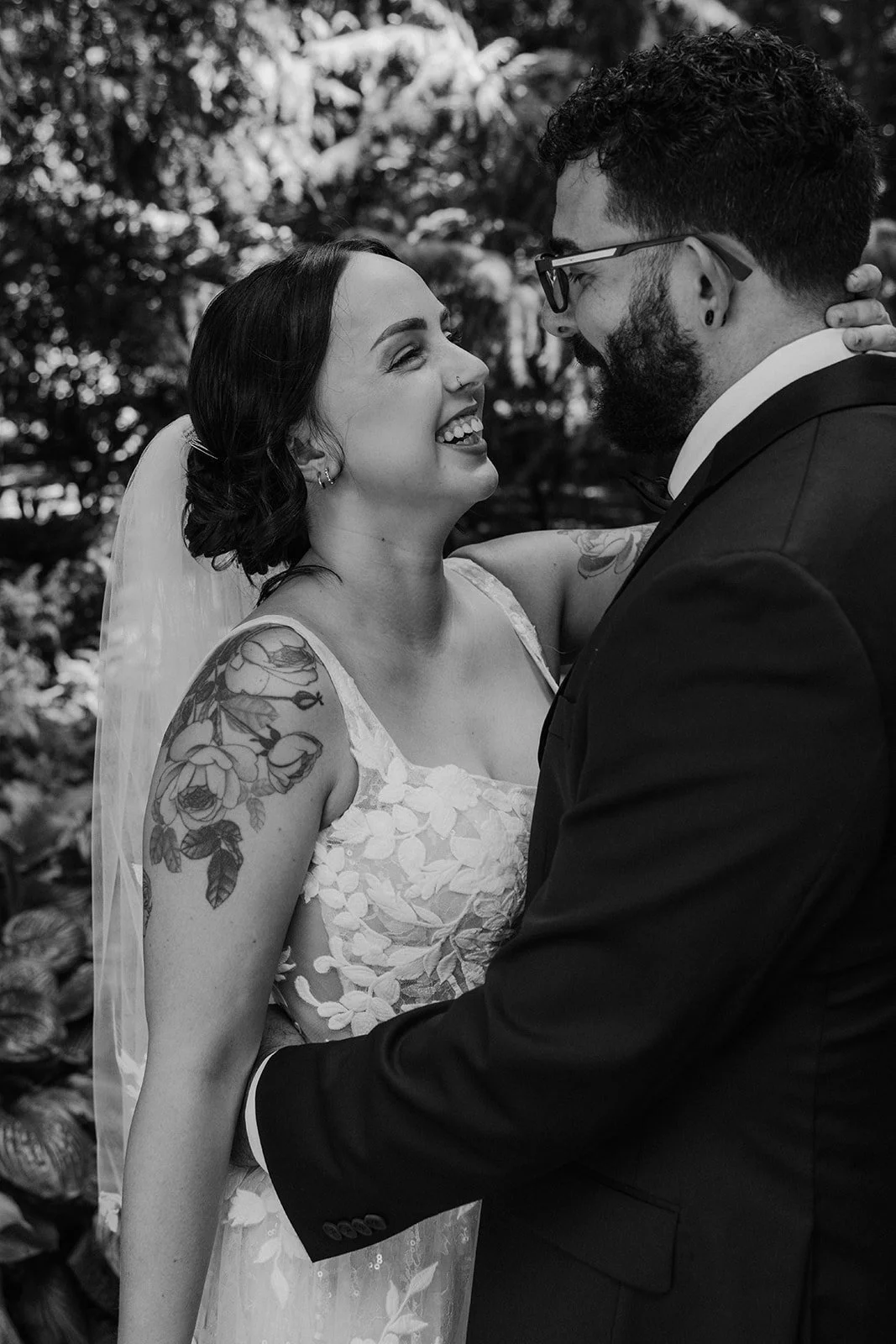 A black and white photo of a bride and groom smiling and embracing, with the bride showing a tattoo on her shoulder, and the groom wearing glasses.