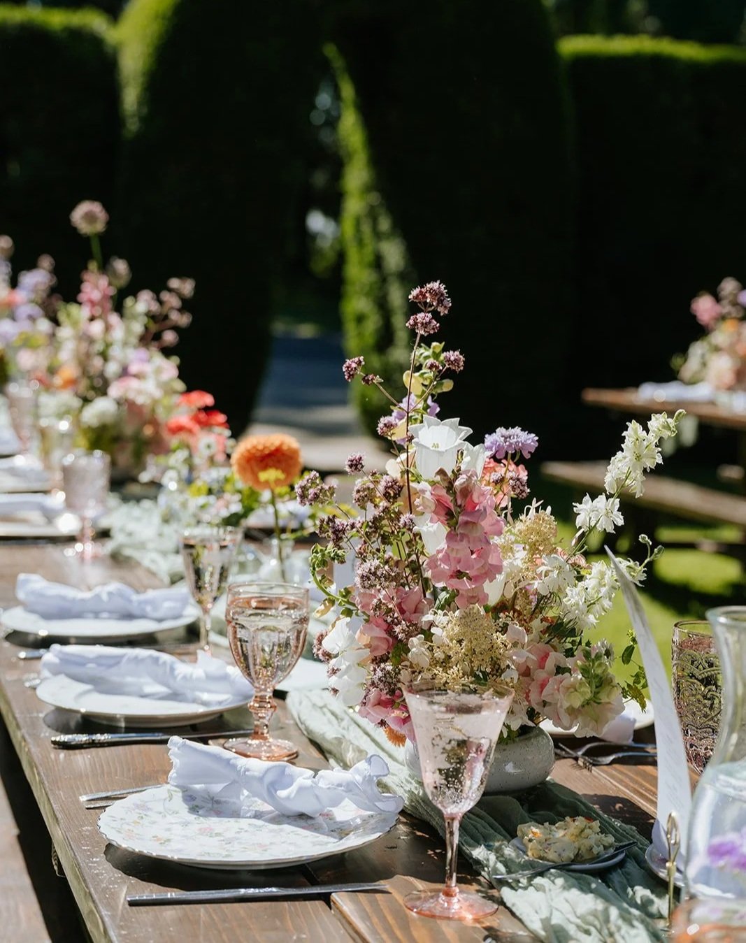 Elegant outdoor tablesetting with floral centerpieces, pink and clear glassware, white napkins, and plates, set in a lush garden with green hedges. Humble Bouquet, Evergreen Gardens, Ferndale Washington
