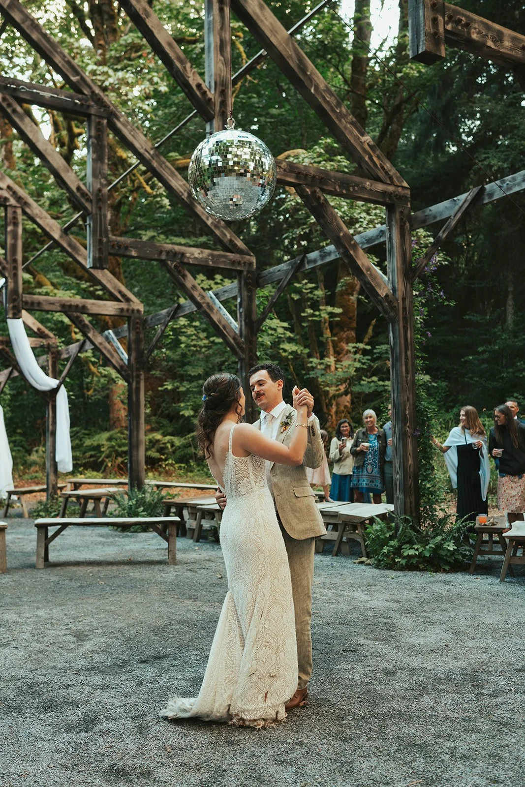 A bride and groom dance outdoors at their wedding reception in a wooded area under a wooden pergola with a disco ball hanging above them. Guests watch in the background.