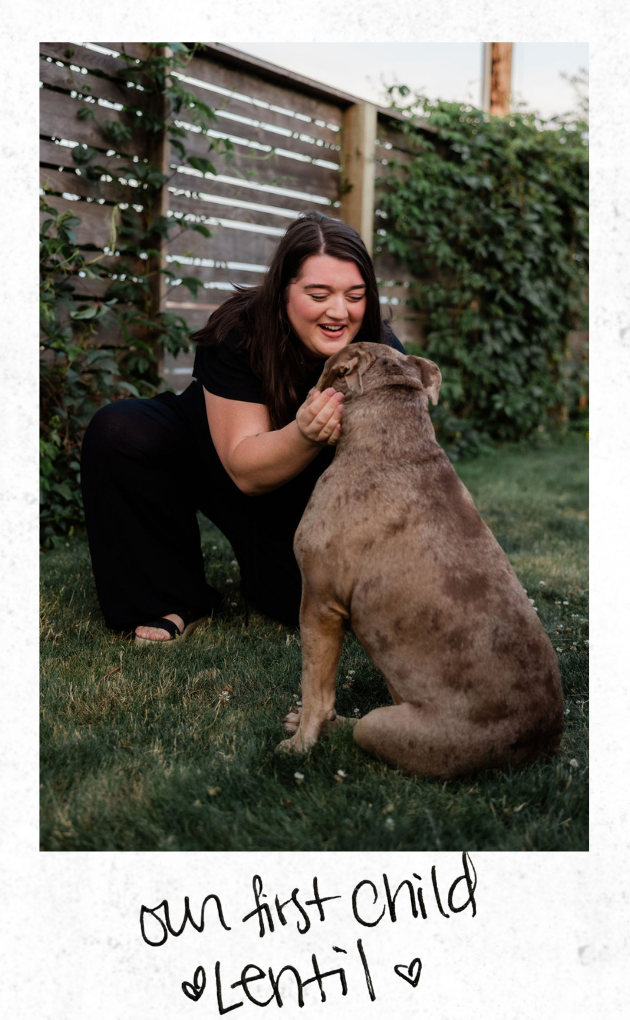 A woman with dark hair smiling and petting an old english bulldog, in a backyard with a wooden fence and greenery in the background.