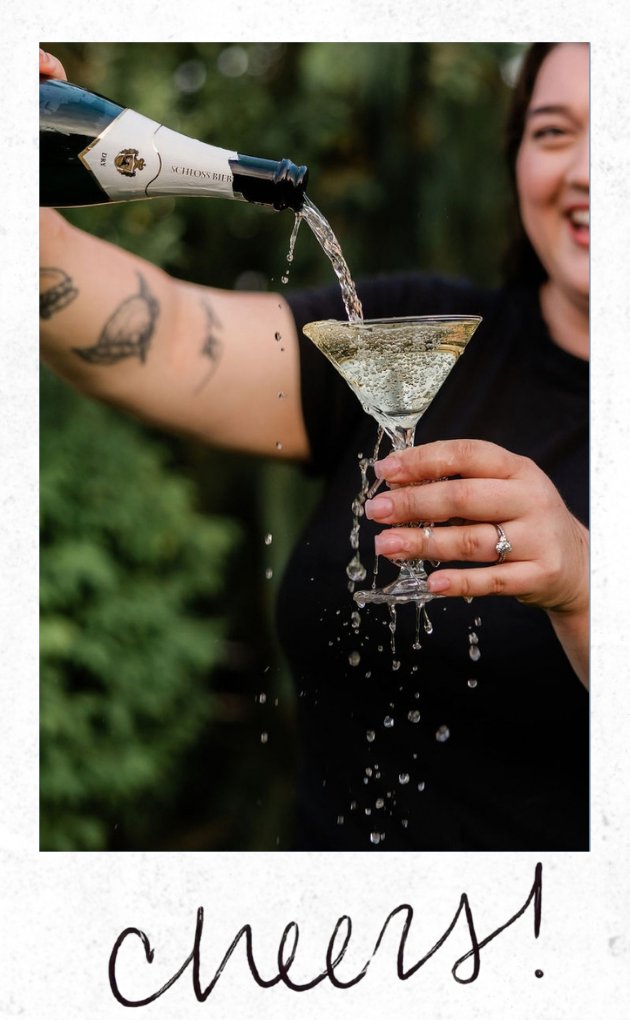Person pouring champagne into a martini glass outdoors, smiling with tattoos visible on arm, the word "cheers!" written at the bottom.