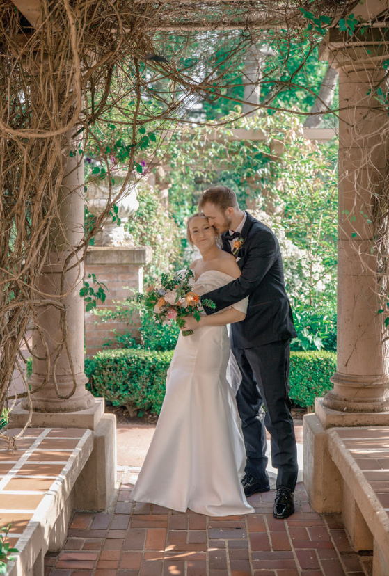 A newlywed couple in wedding attire standing closely and embracing in a garden with greenery and columns. Lairmont Manor, Bellingham Washington