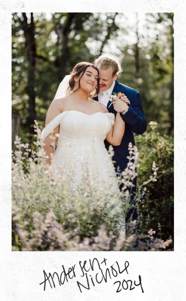 A bride and groom sharing a joyful moment outdoors, surrounded by greenery and purple flowers. The bride wears a white wedding dress and the groom a navy blue suit.