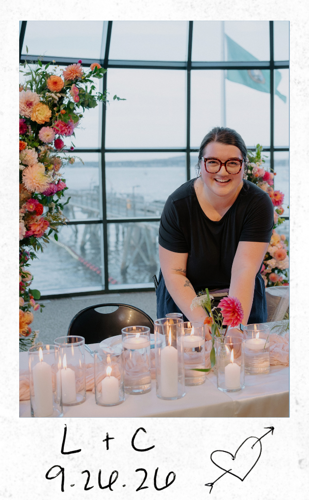 A woman with dark hair, glasses, and tattoos, smiling at a table decorated with candles, flowers, and a pink cloth, inside an airport with large windows and a plane visible outside. Handwritten text and heart drawing are on the bottom of the photo.