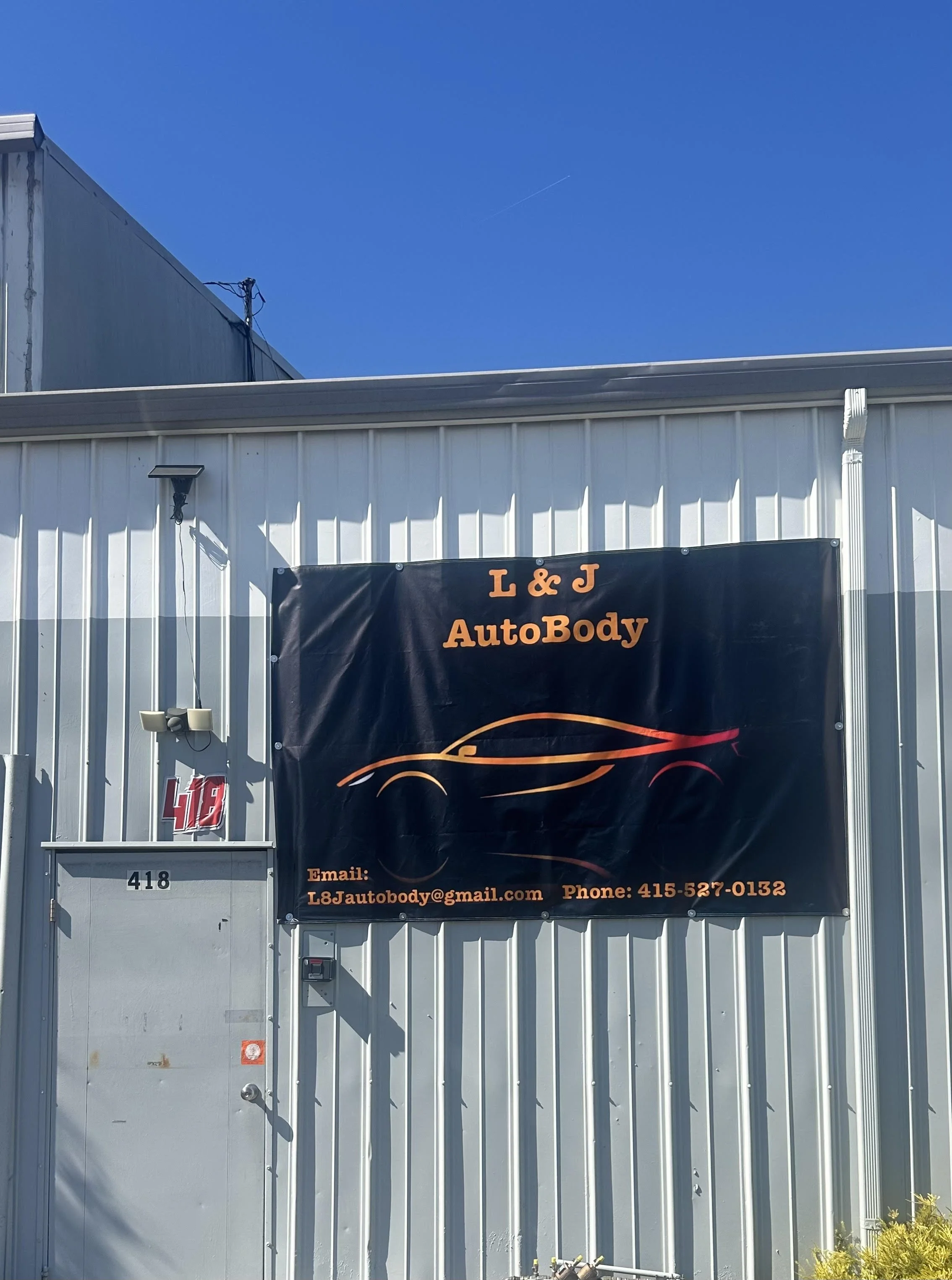 The exterior of a building with a banner for L & J AutoBody, including contact information. The building has a white metal siding, a door with the number 418, and a security camera.