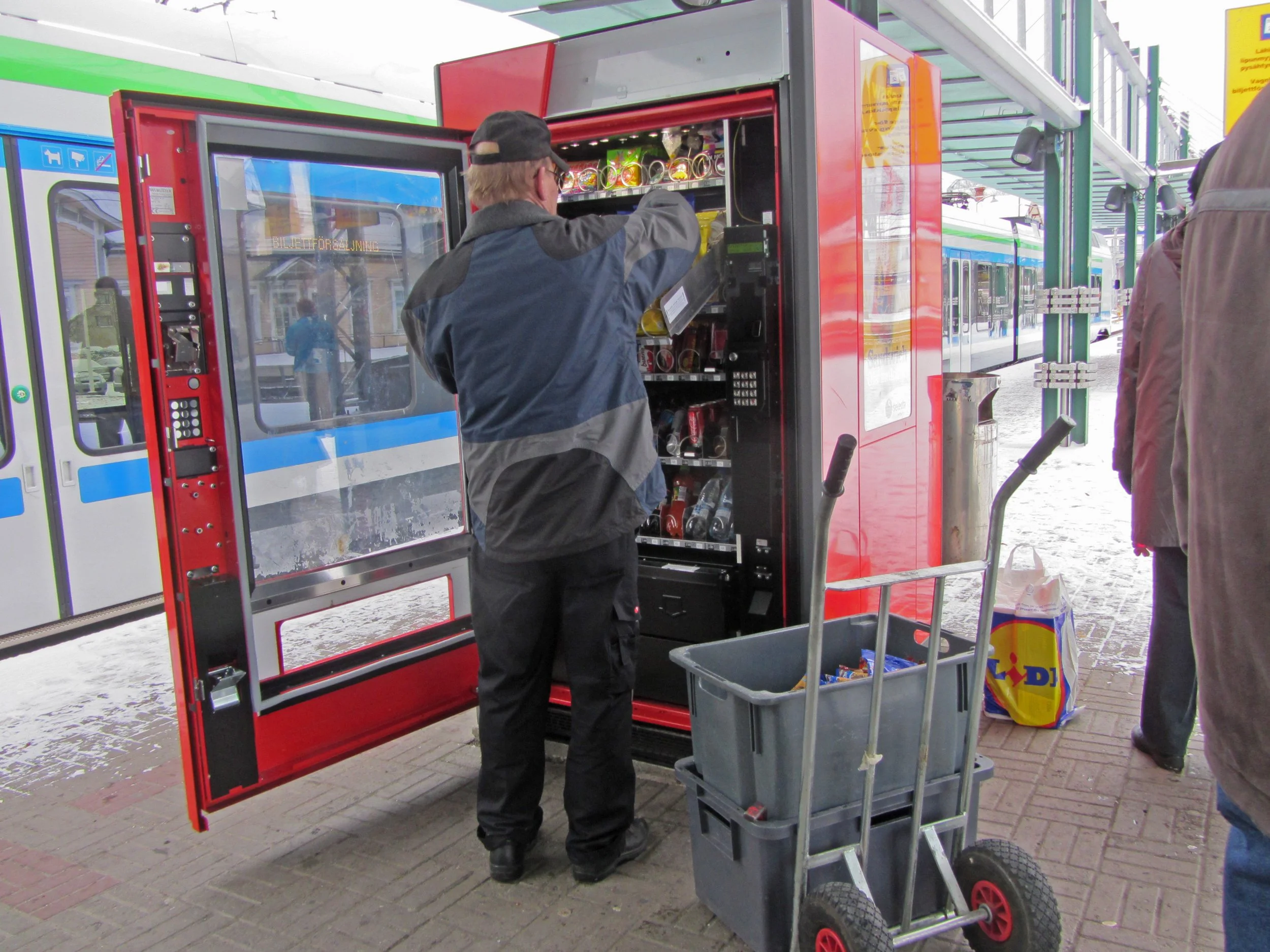 A man in a gray jacket, black cap, and glasses is operating a red vending machine on a city sidewalk. The vending machine contains snacks or drinks, with part of a shopping cart filled with a Lidl shopping bag nearby. Other people are visible in the background near train platforms.