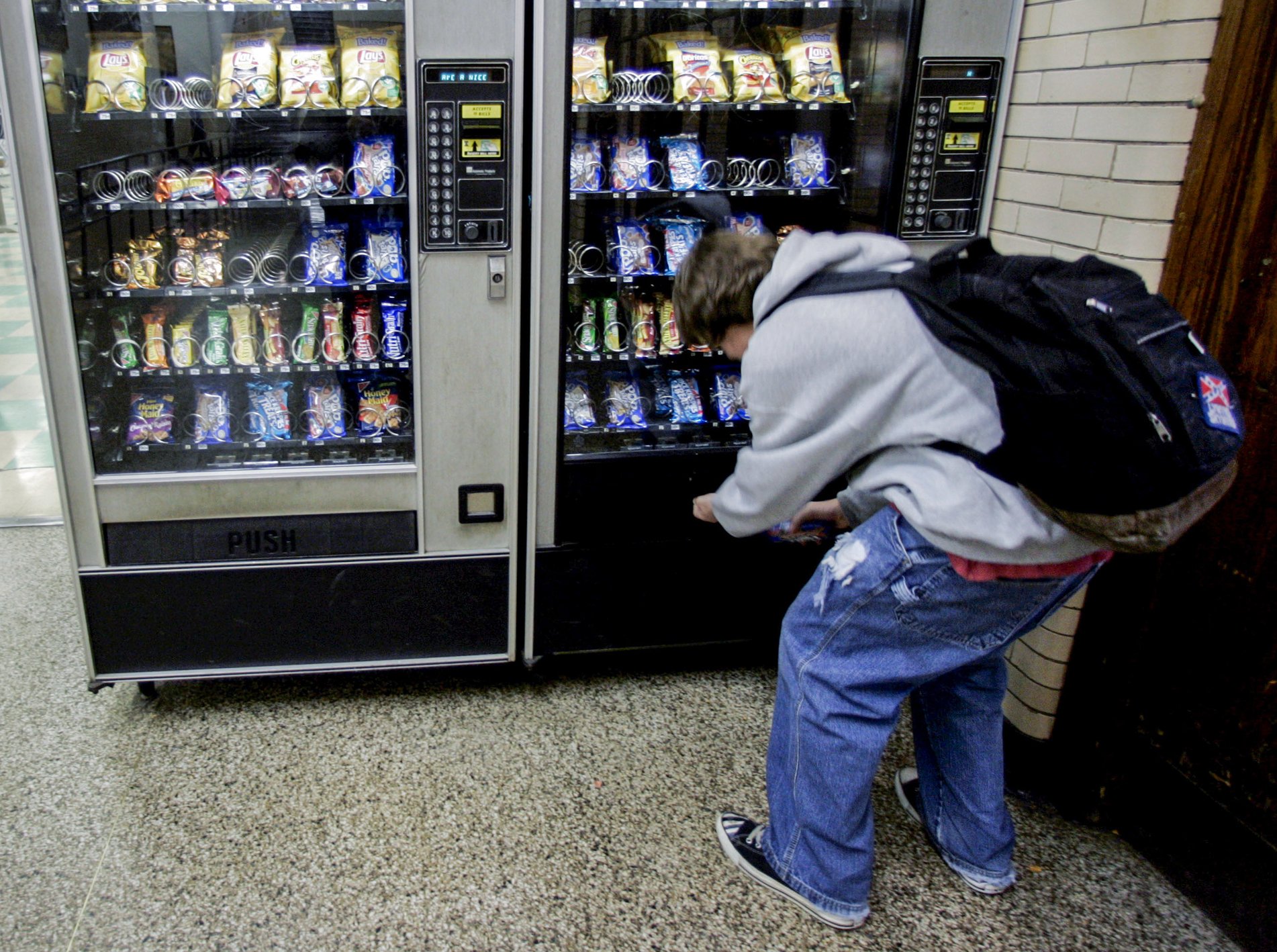 A person crouching in front of a vending machine filled with snacks, with a backpack and wearing blue jeans and a gray hoodie.