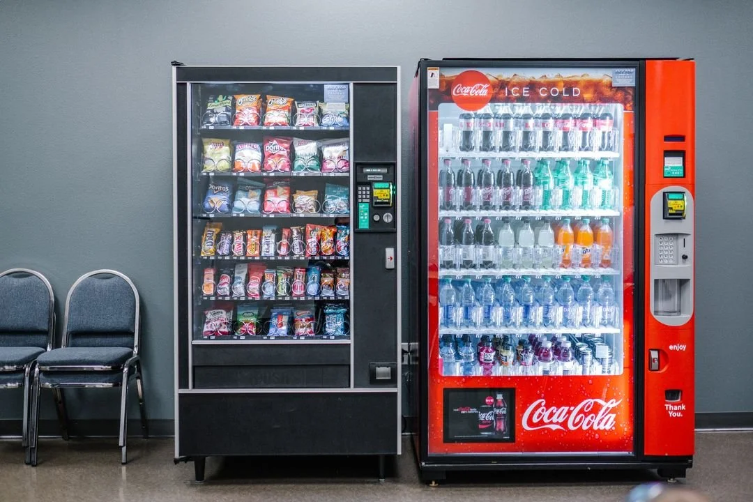 A vending machine with snack options on the left and a Coca-Cola vending machine on the right against a plain wall, with two empty chairs to the left.