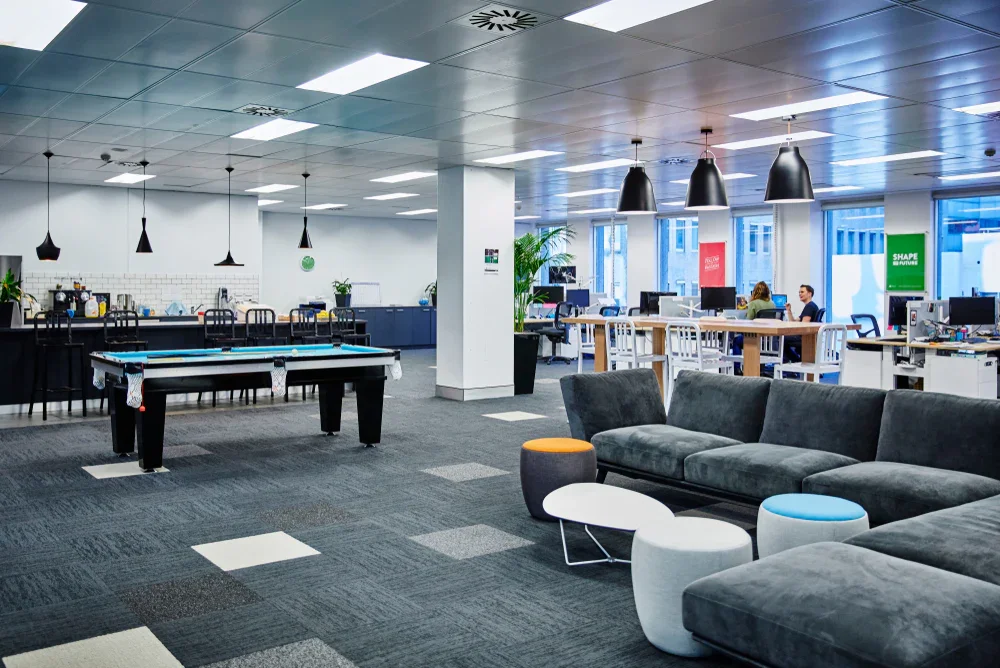 Modern open-plan office space with a lounge area, pool table, and workstations, illuminated by hanging pendant lights and large windows.