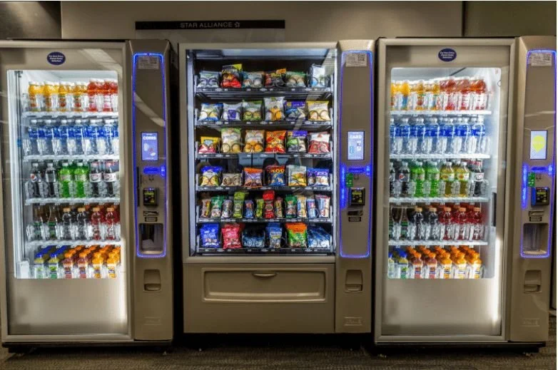 Vending machines filled with snacks and beverages, including chips, candy, and drinks, in an airport or convenience store setting.
