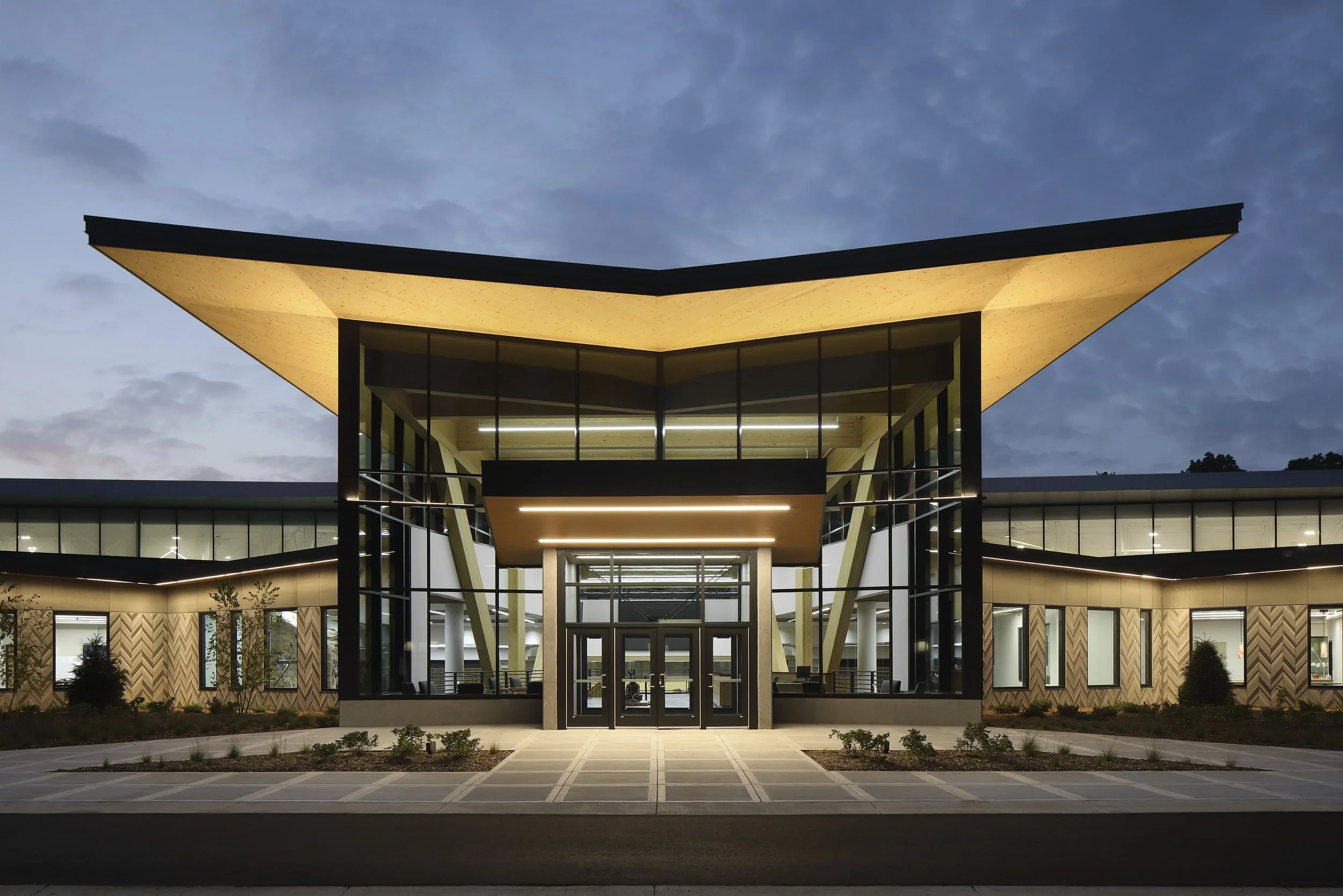 Modern building with a unique upwards V-shaped roof, illuminated from below, during twilight, with large glass windows and a landscaped foreground.