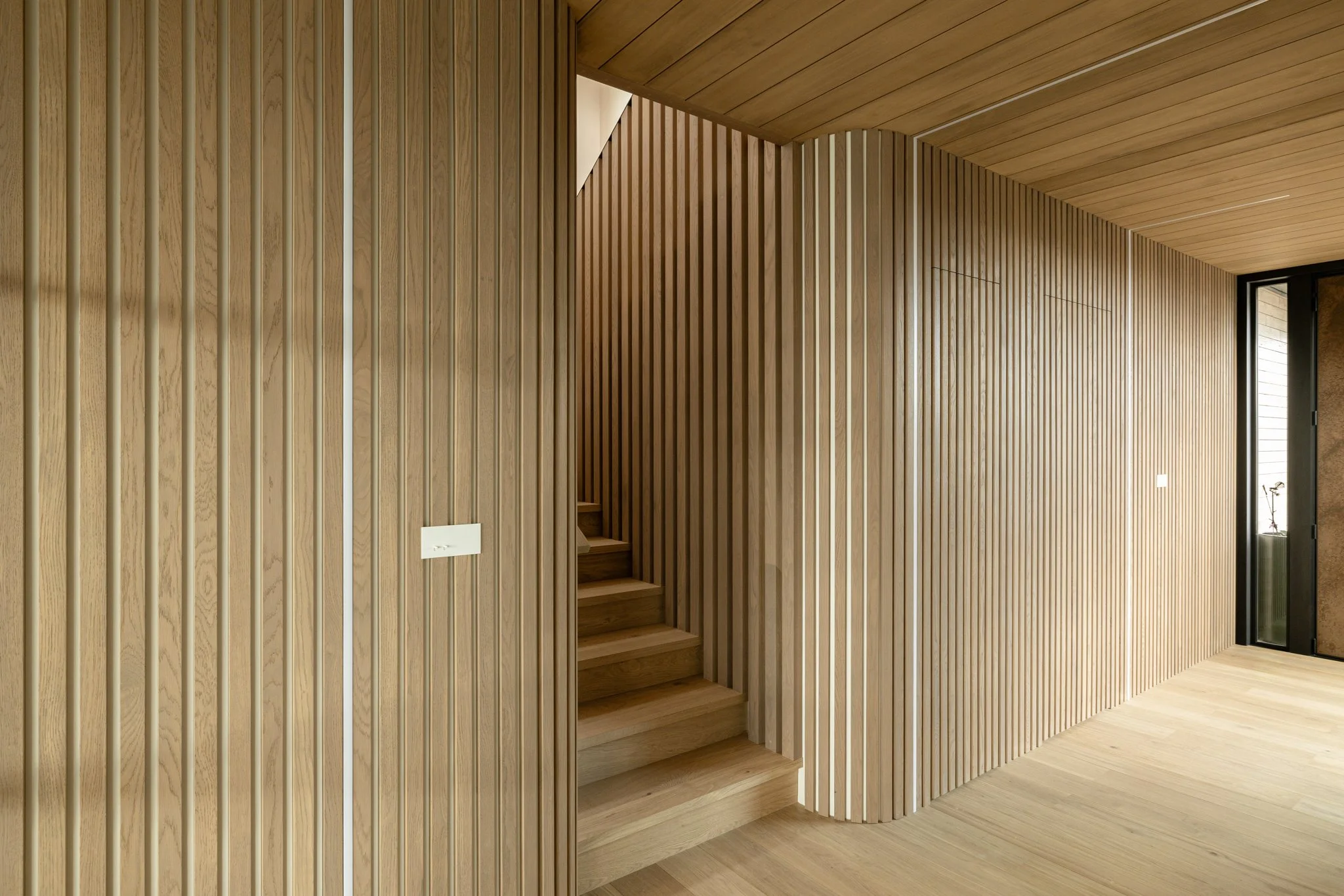 Interior of a modern home with vertical wooden slat walls, a small staircase, and a large window with a door leading outside.