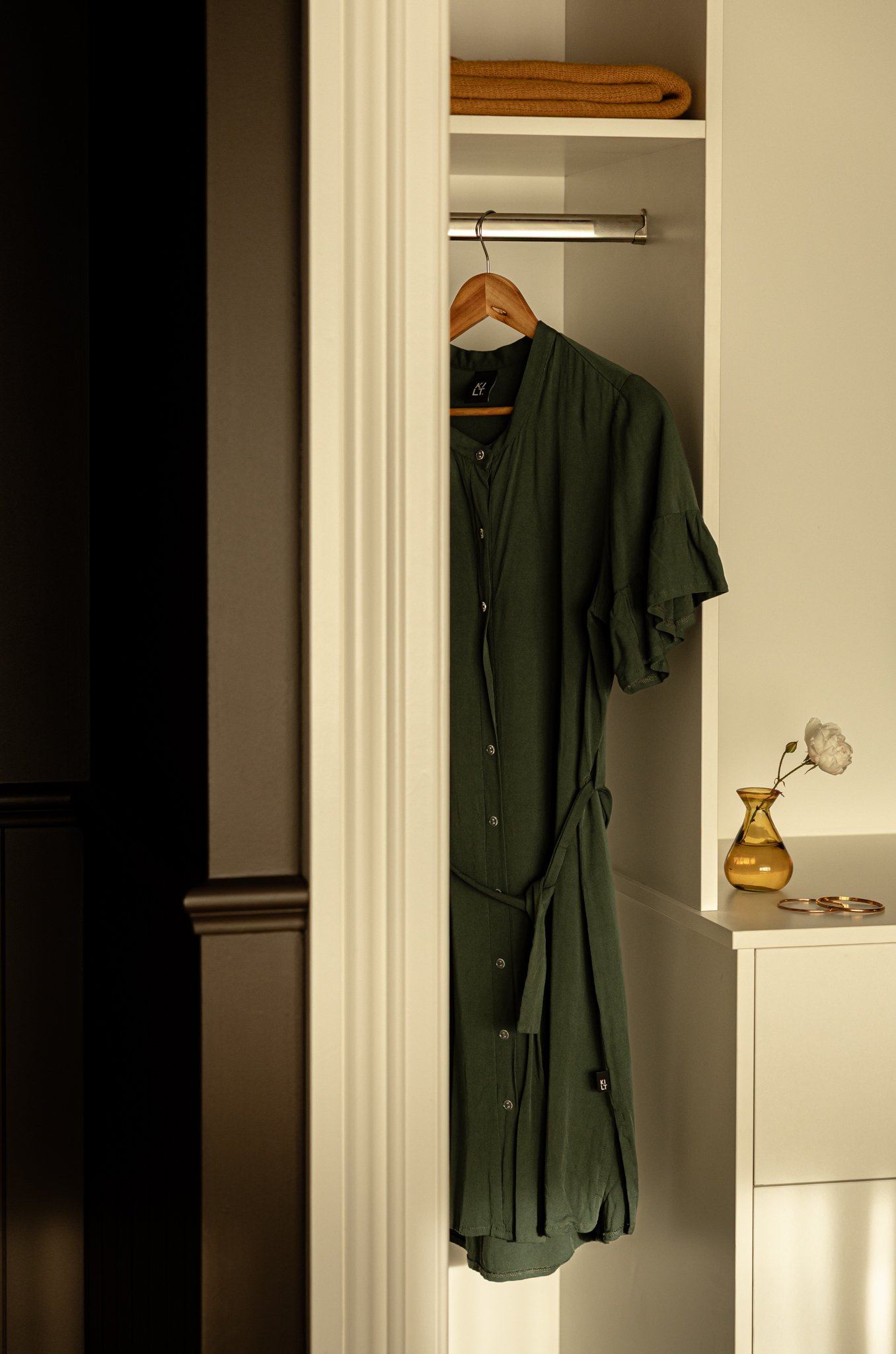 Inside a white wardrobe, a green coat hangs on a wooden hanger on a metal rod. On the top shelf, there are folded tan towels. To the right, a white nightstand with a gold vase holding a flower and a pair of gold earrings.