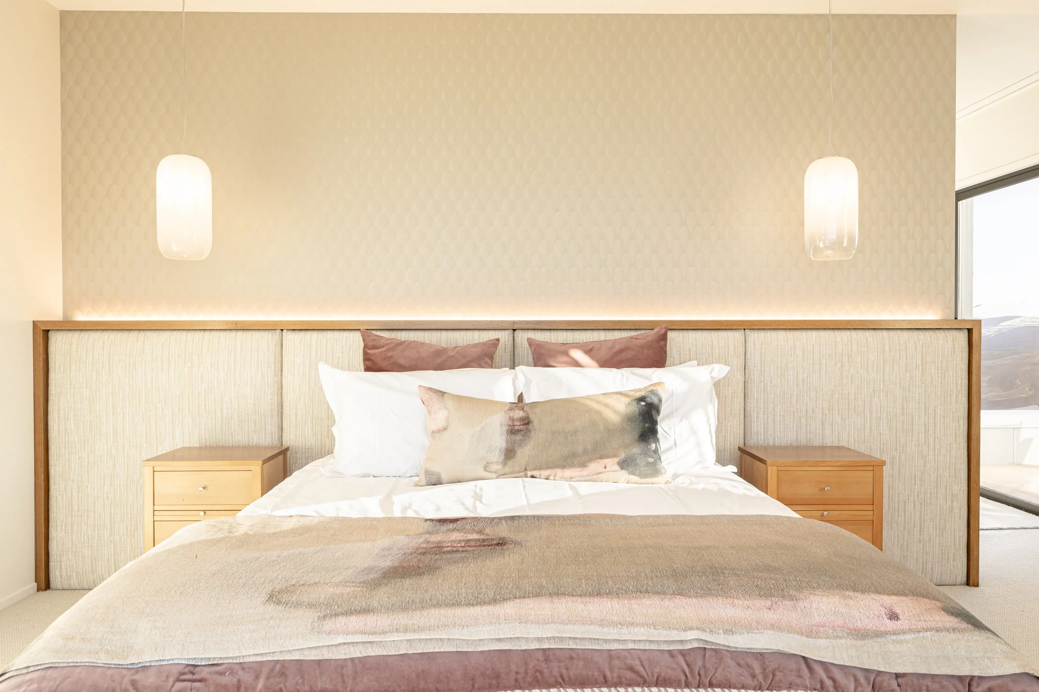 A tidy bedroom with a large bed, brown and white pillows, two wooden nightstands, and modern hanging pendant lights on a textured beige and cream wall.