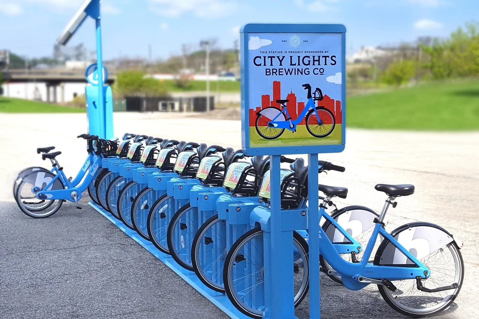 Blue bike-sharing station with multiple bicycles and a sign indicating sponsorship by City Lights Brewing Co.