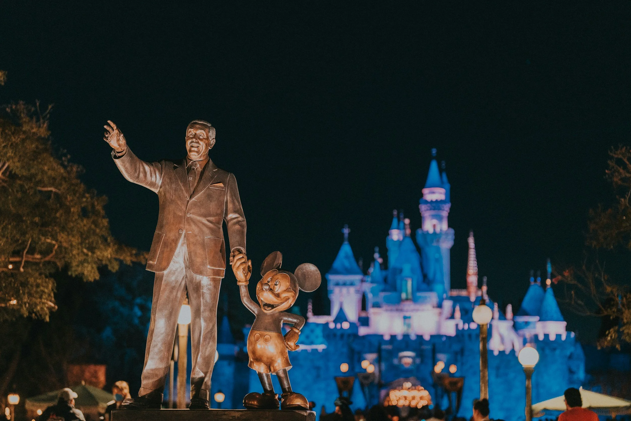 Nighttime view of a bronze statue of Walt Disney with Mickey Mouse and a castle in the background at Disneyland.