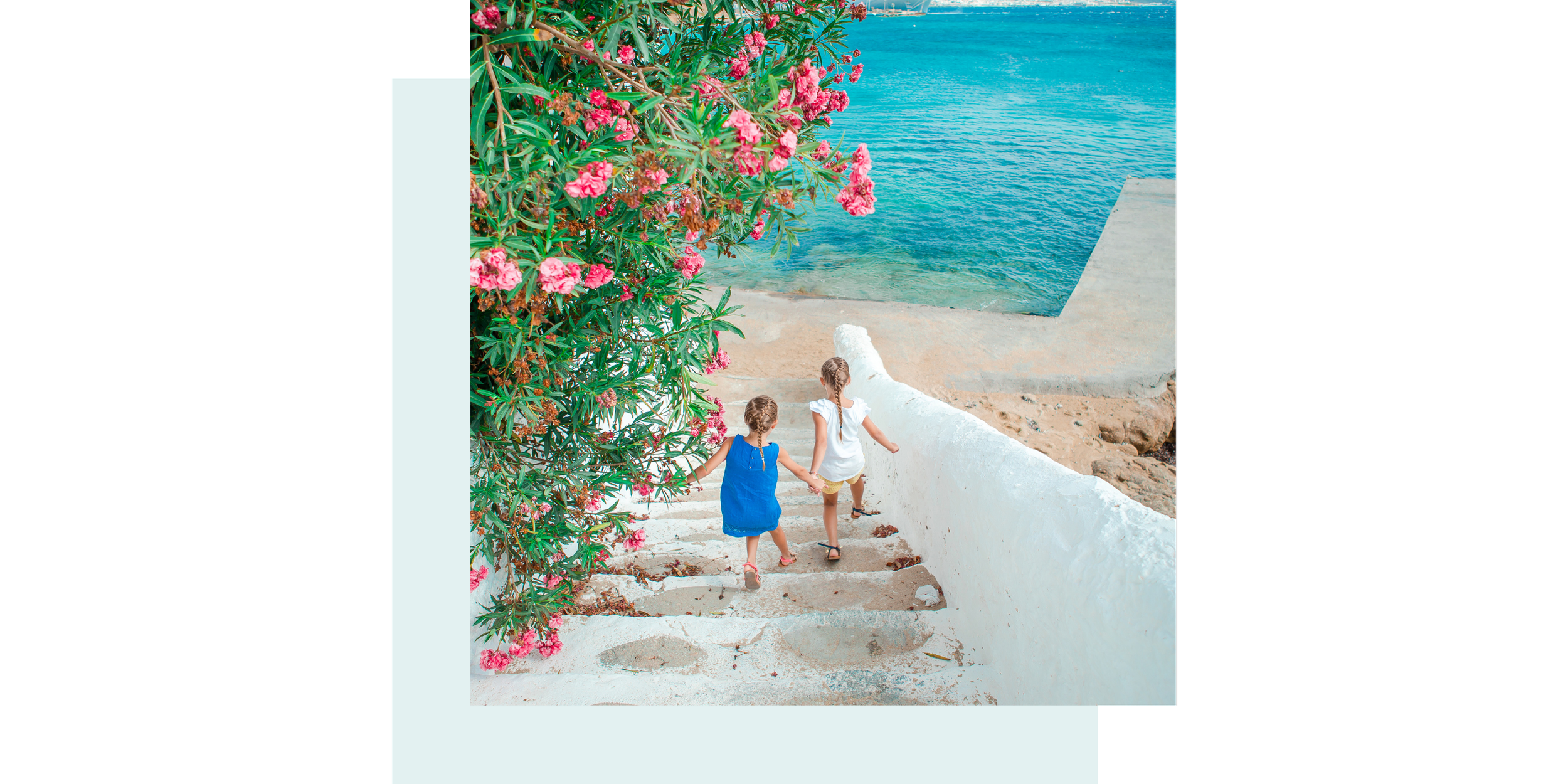 Two young girls with braided hair, one in a blue dress and the other in a white dress, holding hands and walking down white stairs in the Mediterranean, with pink flowers and green bushes on the side, and the ocean in the background.