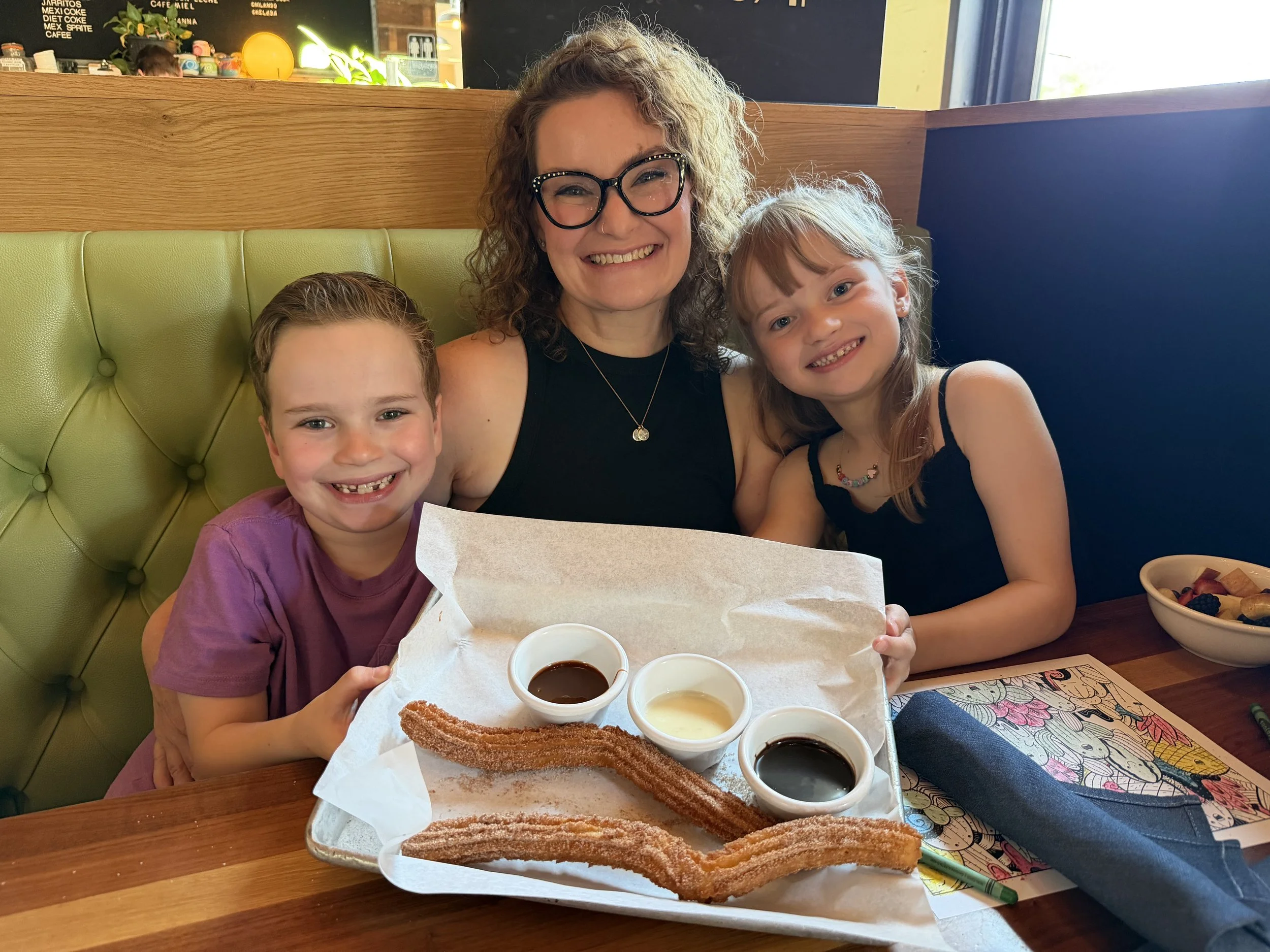 A woman and two children smiling and sitting at a restaurant table. On the table, there is a tray of churros and several small bowls of dipping sauces. The background shows a restaurant interior with menu boards and a window.