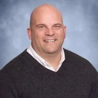 Portrait of a bald man with a goatee, wearing a white shirt and a black sweater, smiling against a blue background.