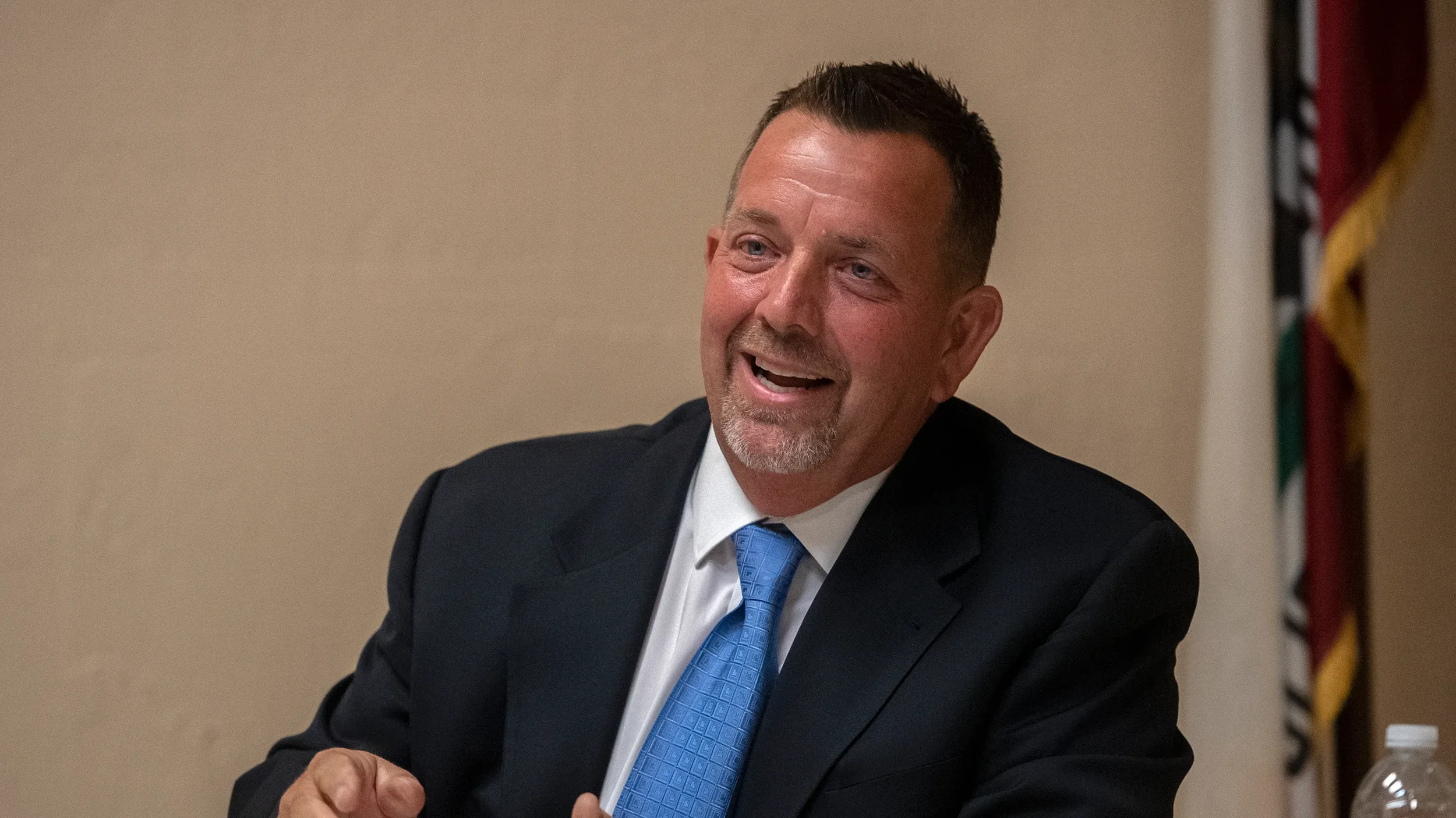 A man in a black suit, white shirt, and blue tie sitting at a table, smiling and speaking, with a flag partially visible in the background.