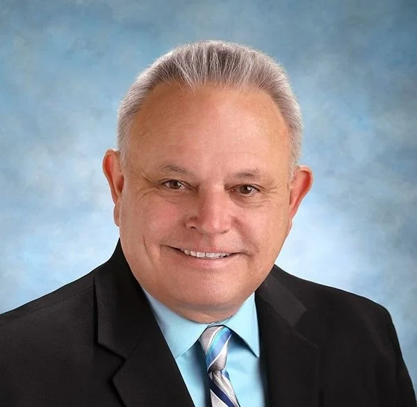 A middle-aged man with short gray hair, smiling, wearing a dark suit, light blue shirt, and a striped tie, against a blue background.