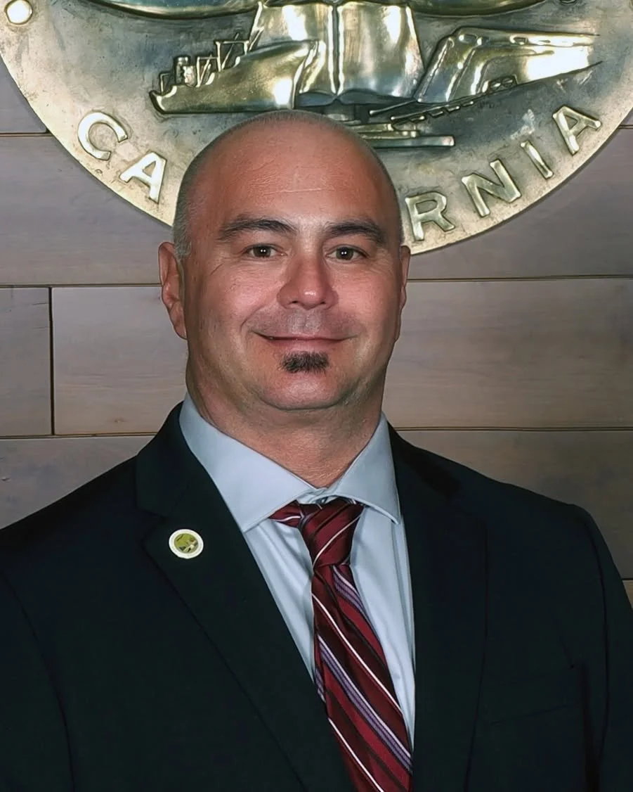 A man in a black suit, white shirt, and red striped tie smiling, standing in front of a wall with a metal emblem that has a ship and the words "California" and "California" partially visible.