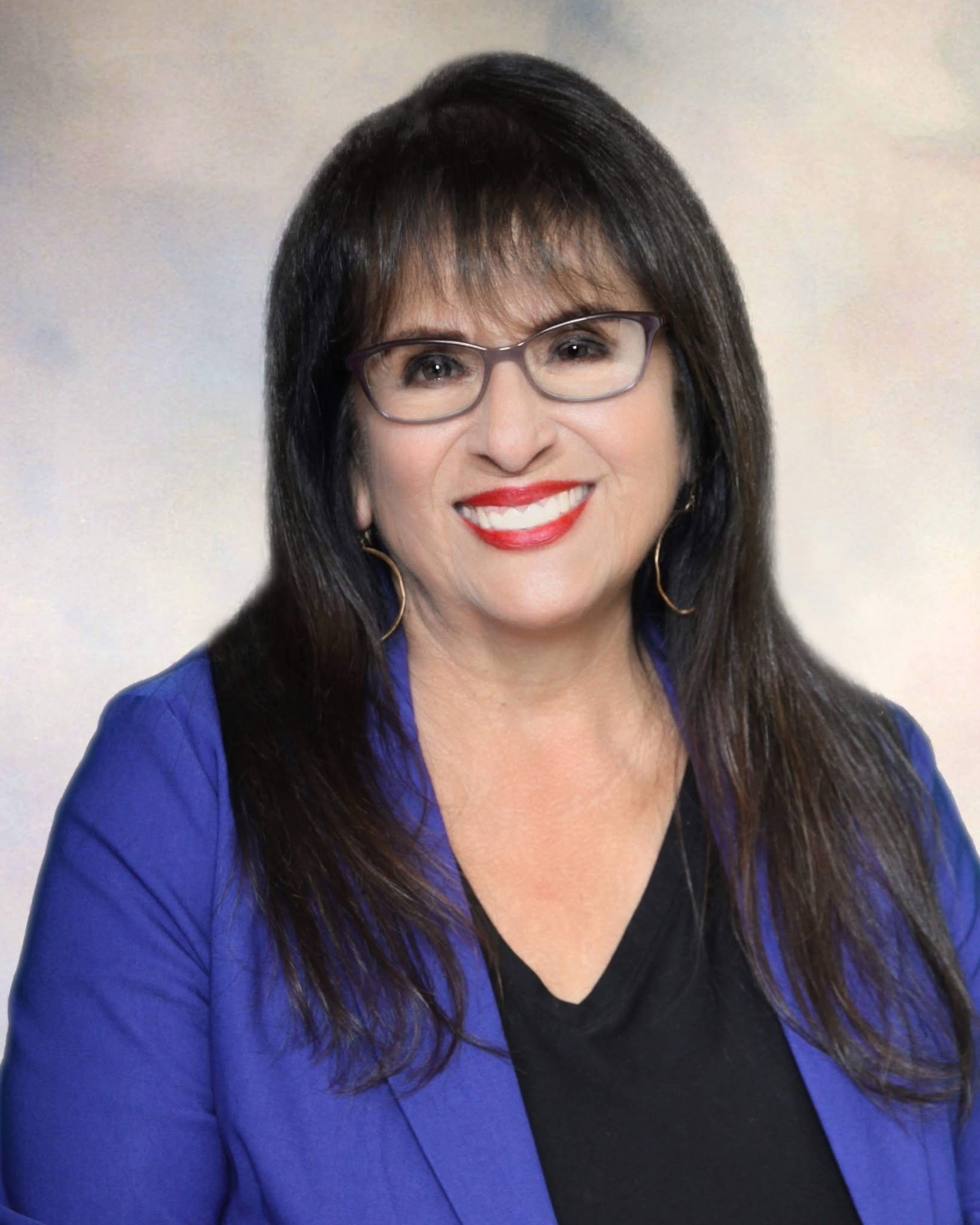 A woman with long dark hair, glasses, red lipstick, and earrings wearing a blue blazer and black top, smiling at the camera.