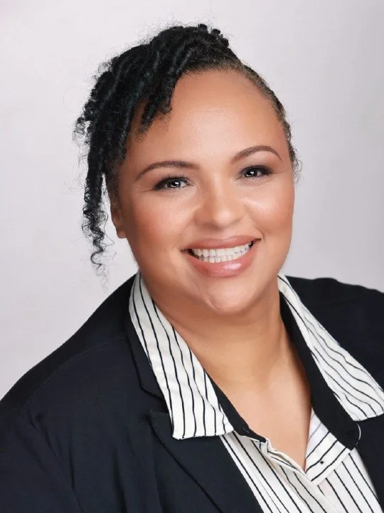 Professional headshot of a woman smiling, with curly hair and wearing a black blazer over a striped shirt, against a light gray background.