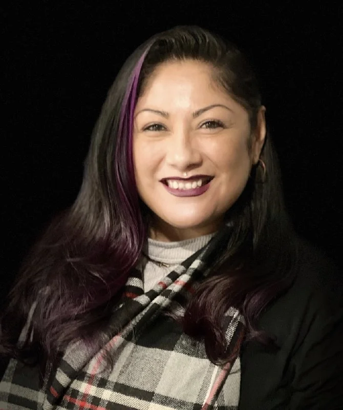 A young woman with dark hair featuring purple streaks, smiling, wearing a black jacket, plaid scarf, and hoop earrings, against a black background.
