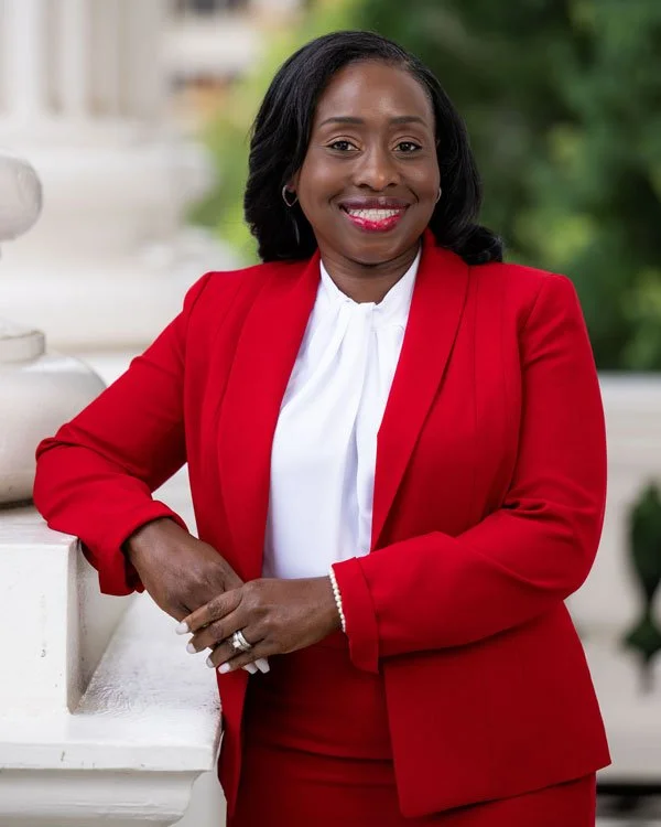 A woman in a red blazer and white blouse smiling outdoors, standing near a white railing with greenery in the background.