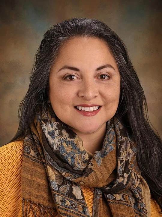 A woman with long dark hair, smiling, wearing a yellow top and a patterned scarf, against a brown gradient background.