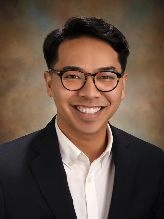A young man wearing glasses, a white dress shirt, and a black blazer, smiling against a blurred brown and beige background.