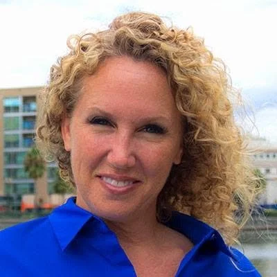 A woman with curly blonde hair wearing a blue shirt outdoors, with buildings in the background.