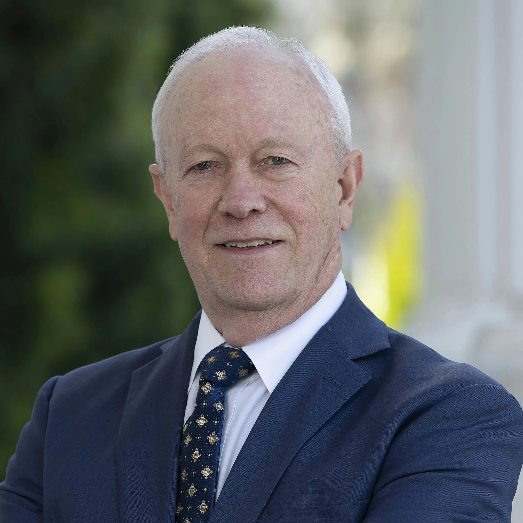 Close-up photo of an elderly man smiling, wearing a dark suit, white shirt, and a patterned tie, with a blurred outdoor background.
