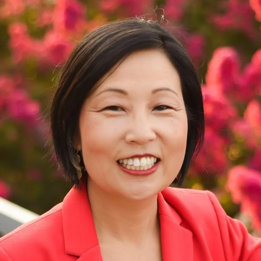 A woman with short black hair smiling with a background of pink and red flowers.
