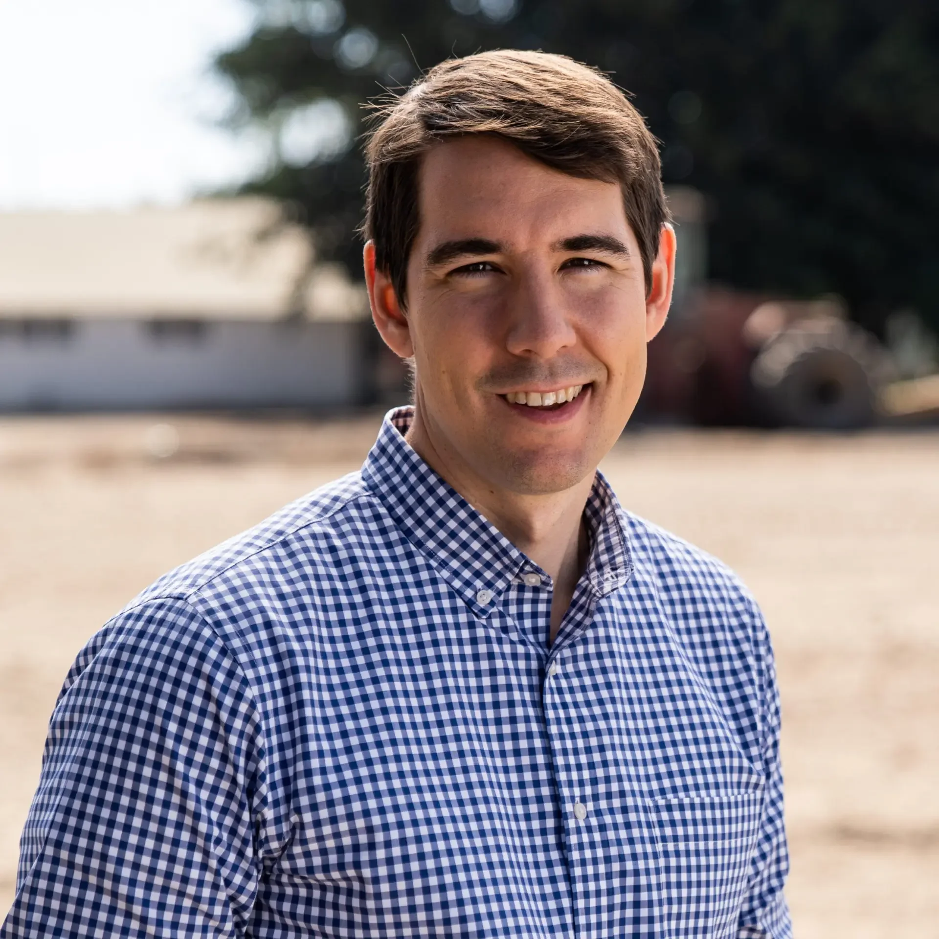 A young man with brown hair smiling outdoors, wearing a blue and white checkered shirt, with a blurred background of trees and buildings.
