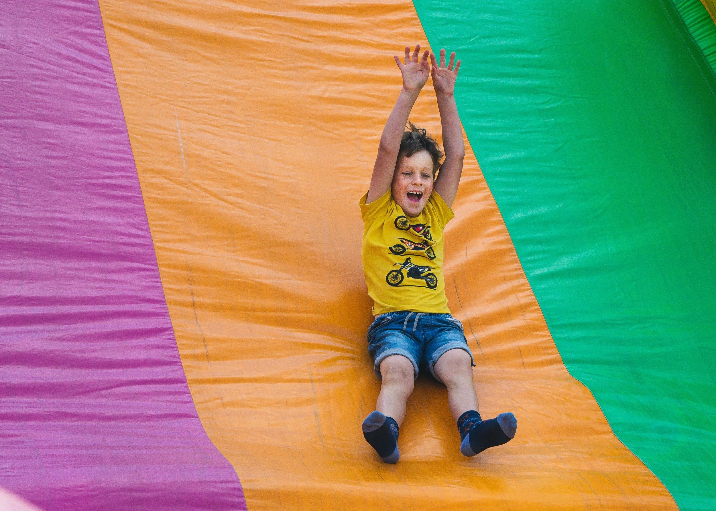 A young boy in a yellow shirt and blue shorts sliding down a colorful inflatable slide with green, orange, and purple sections, with a happy expression and arms raised.