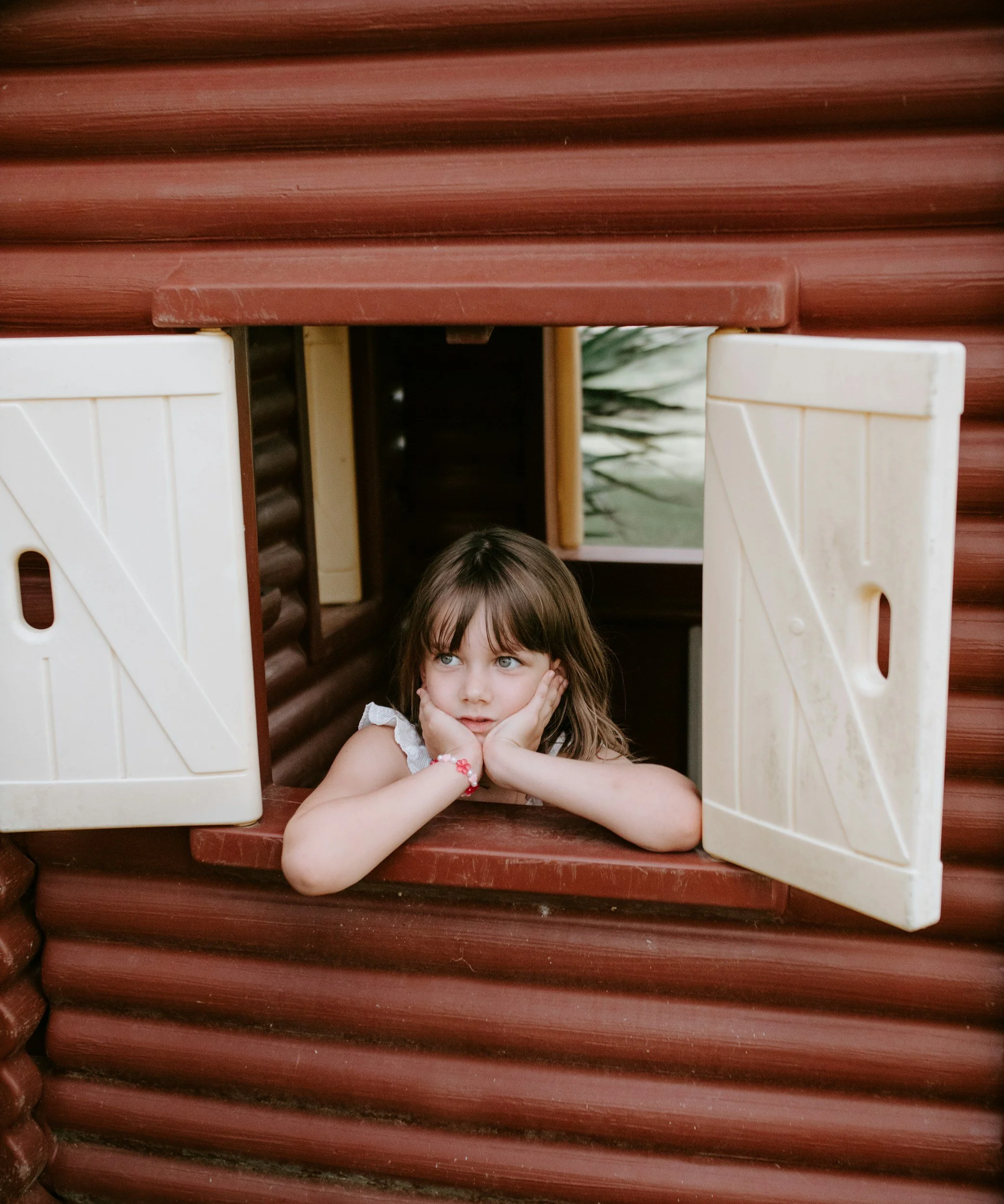 A young girl with brown hair and blue eyes is resting her chin on her hands and looking up through a wooden playhouse window with white shutters.