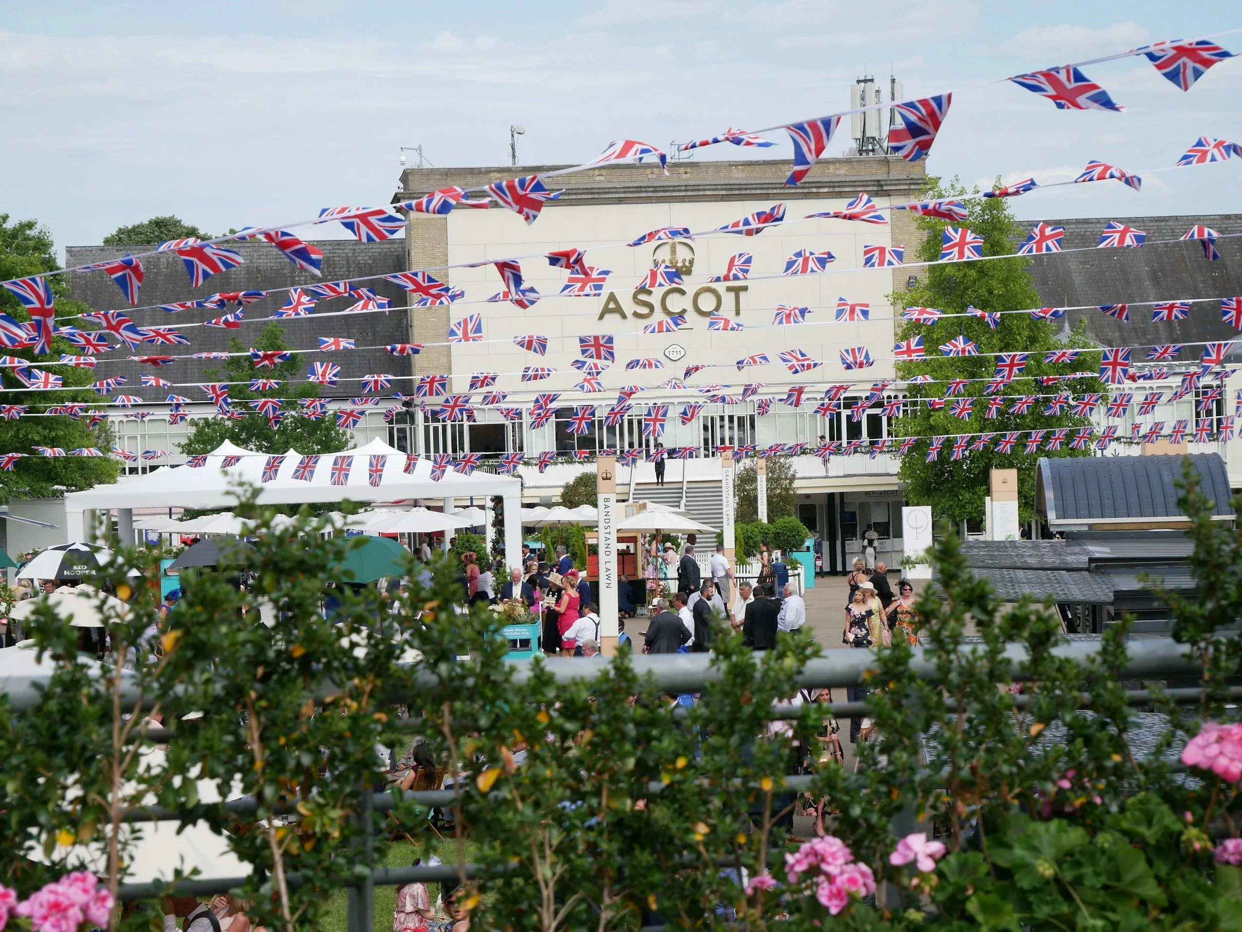 Crowd gathered in front of the Ascot racecourse decorated with numerous Union Jack flags hanging from strings, with some tents and trees visible.